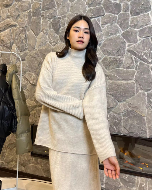 Woman wearing a cream-colored outfit standing against a stone wall.