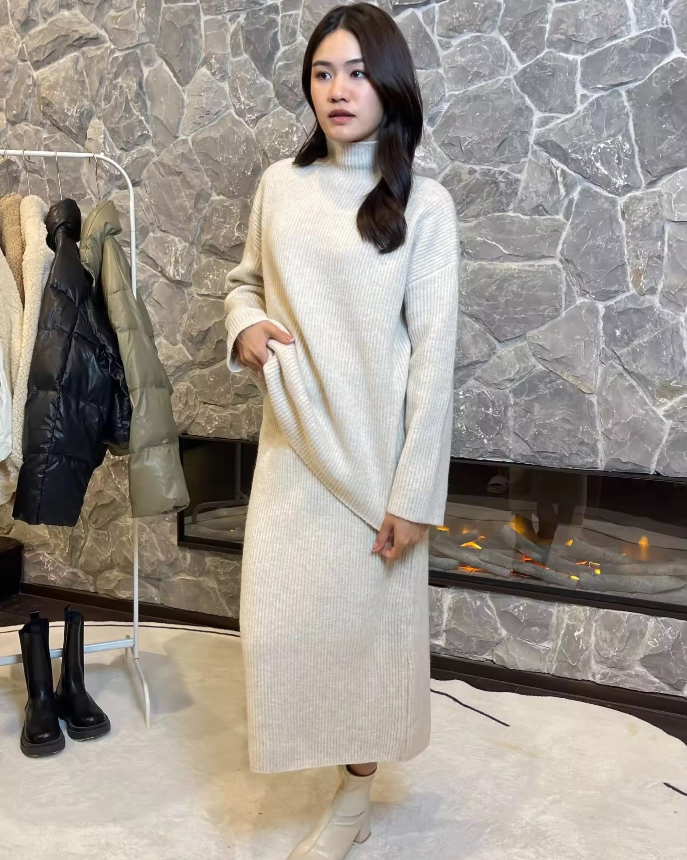 Woman wearing a long white sweater in front of a stone wall.