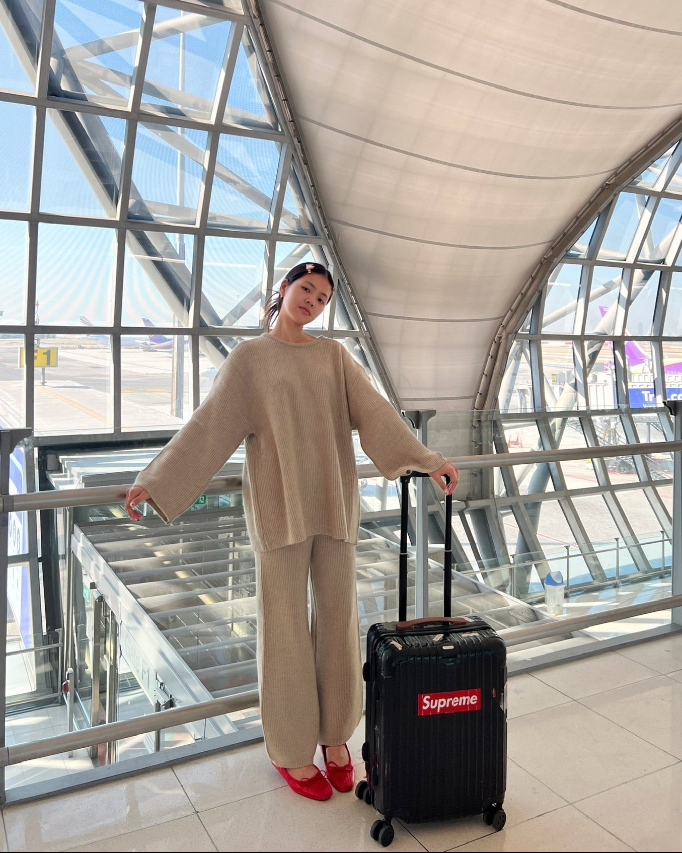 woman in beige outfit with a black suitcase labeled 'Supreme' at an airport terminal.