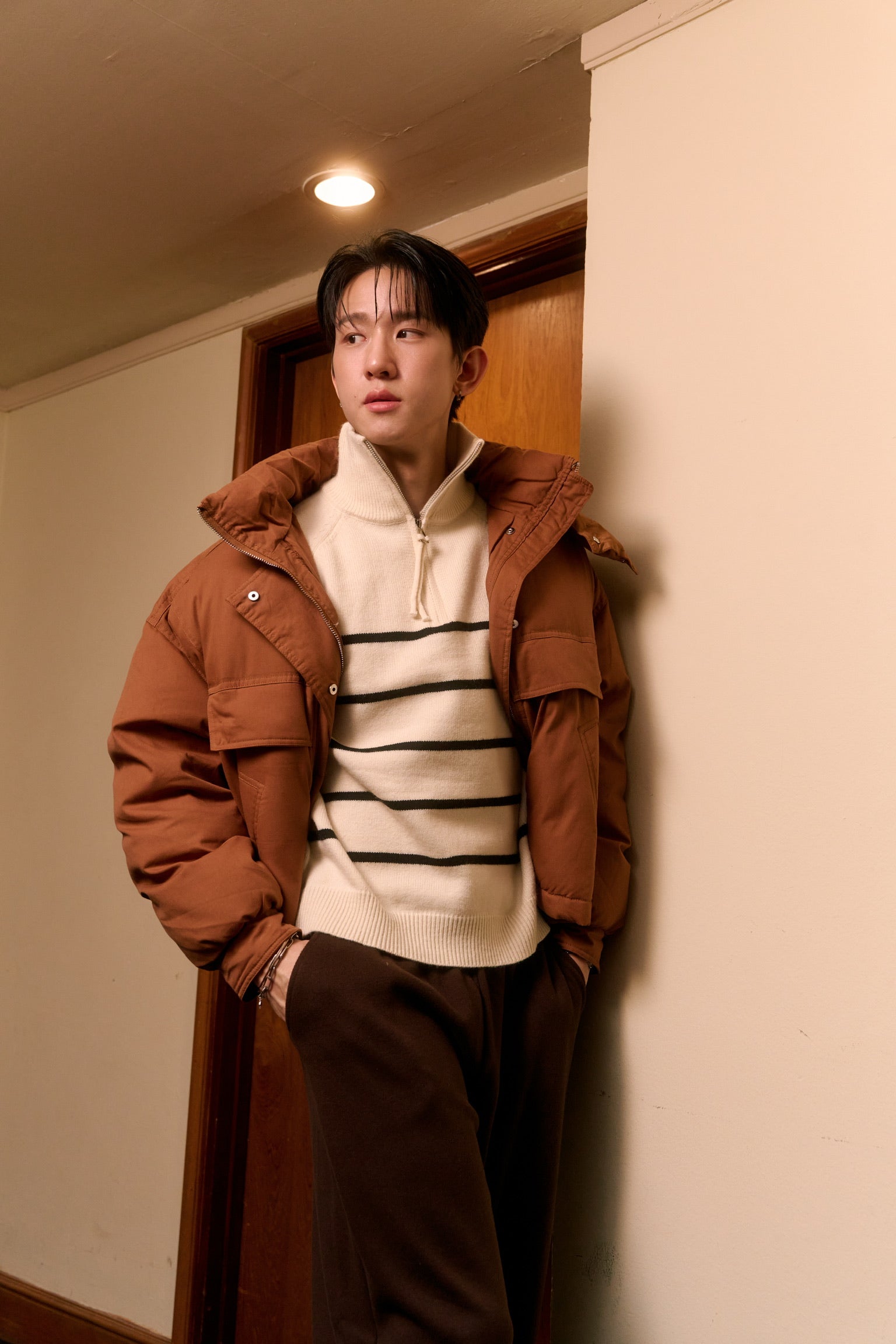 man wearing a brown coat and striped sweater standing against a wall indoors.