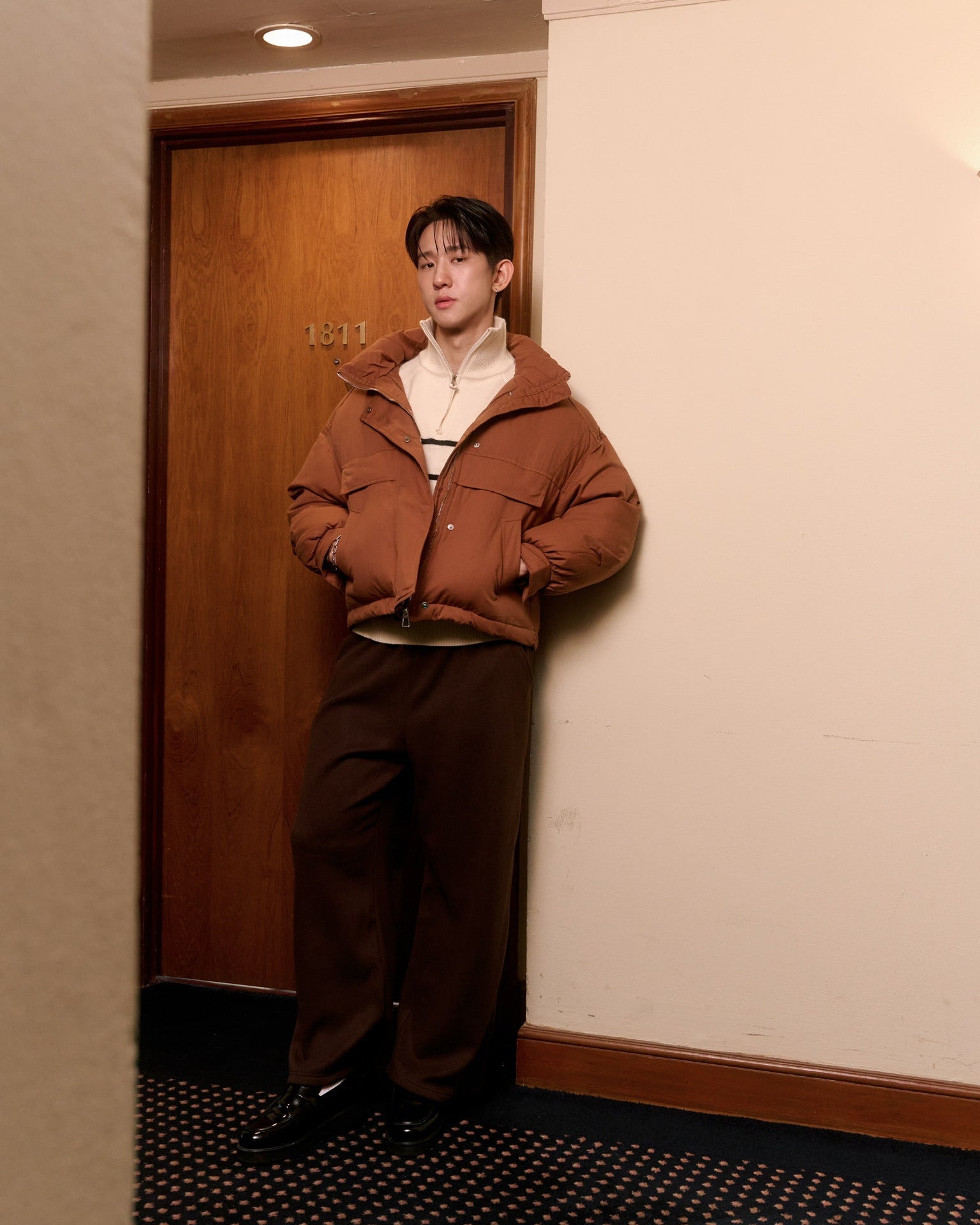 man wearing a brown jacket and pants leaning against a wall.
