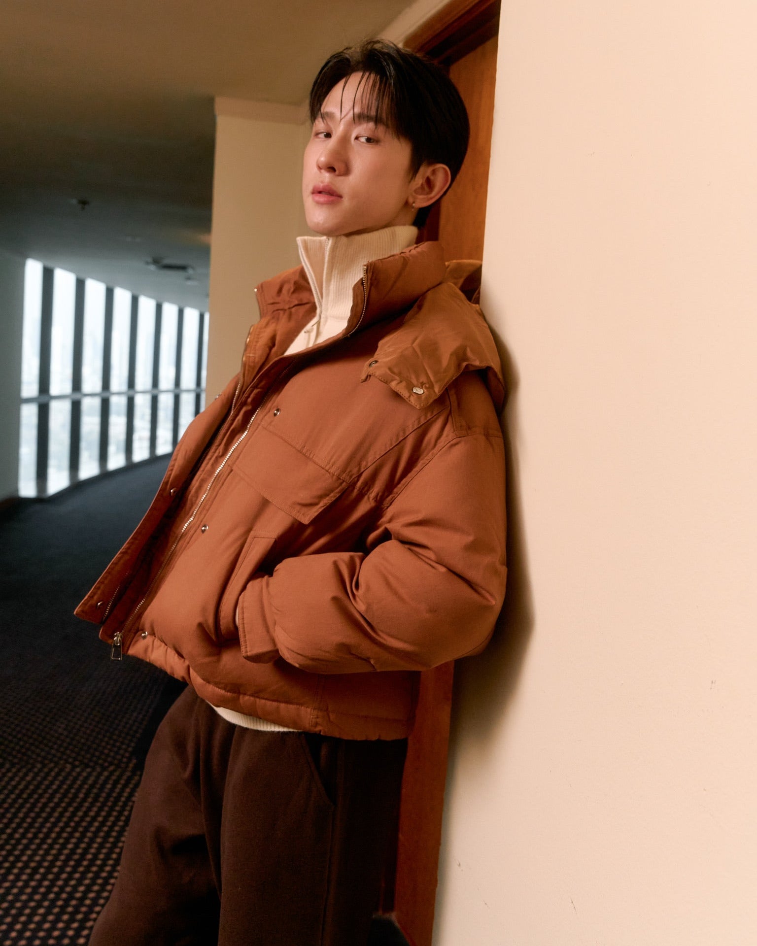 men wearing a brown jacket leaning against a wall indoors
