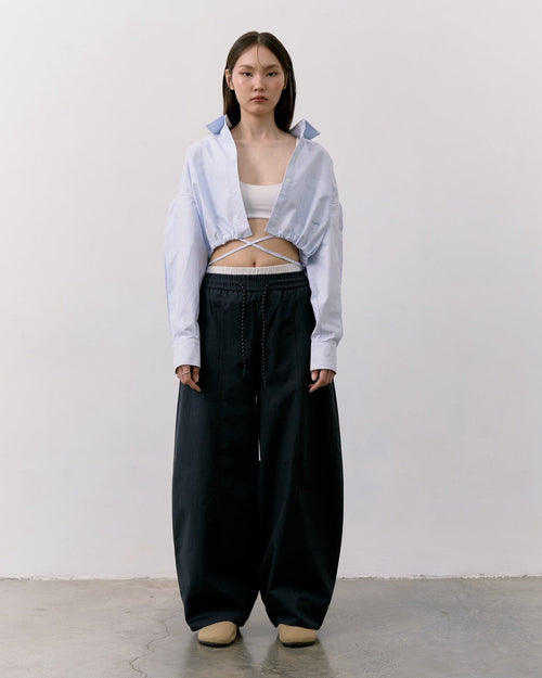 woman stands against a white wall and concrete floor wearing a light blue cropped button-up shirt with a crisscross front detail, loose black wide-leg pants, and beige shoes.