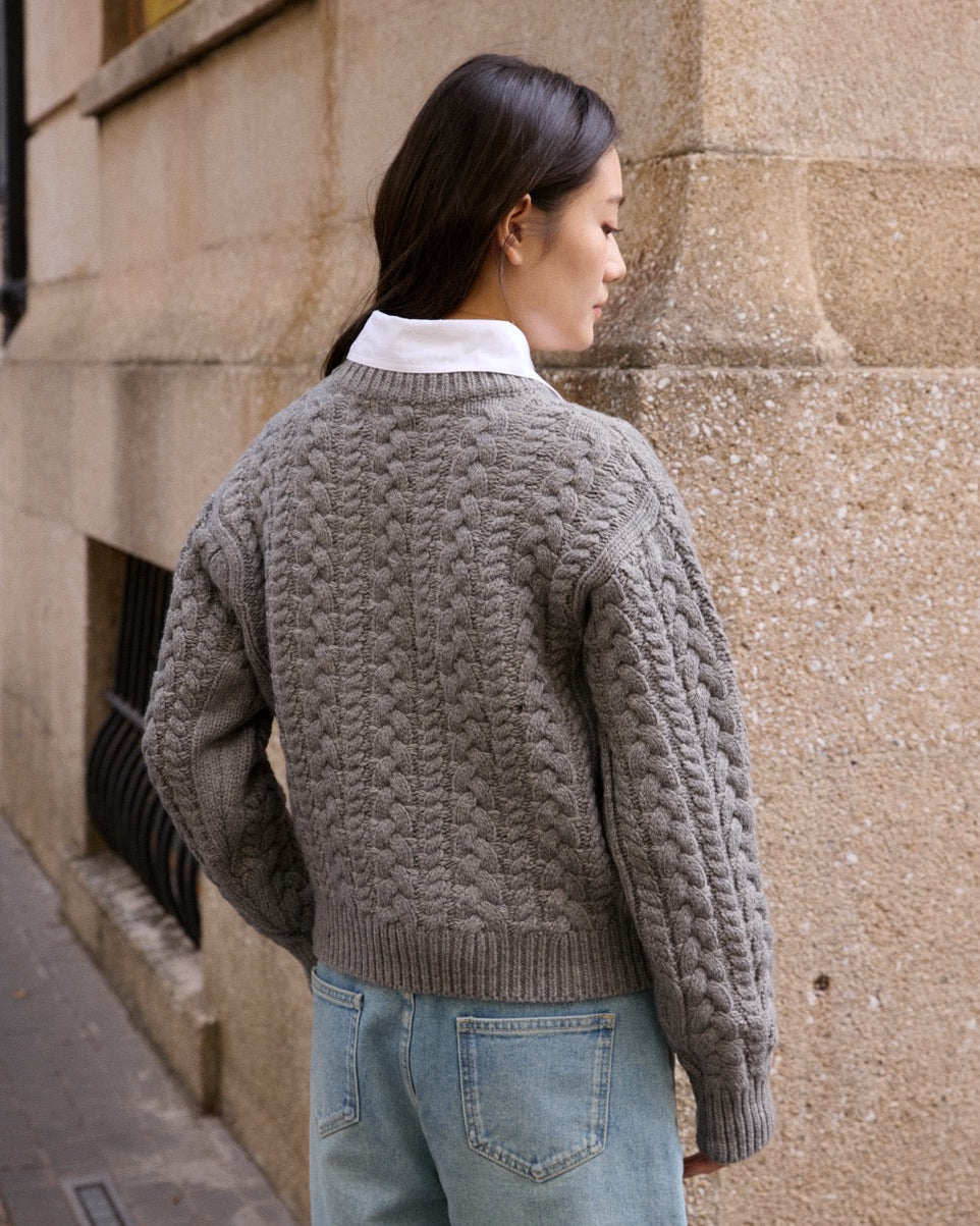 woman wearing a textured gray sweater and light blue jeans against a stone wall.