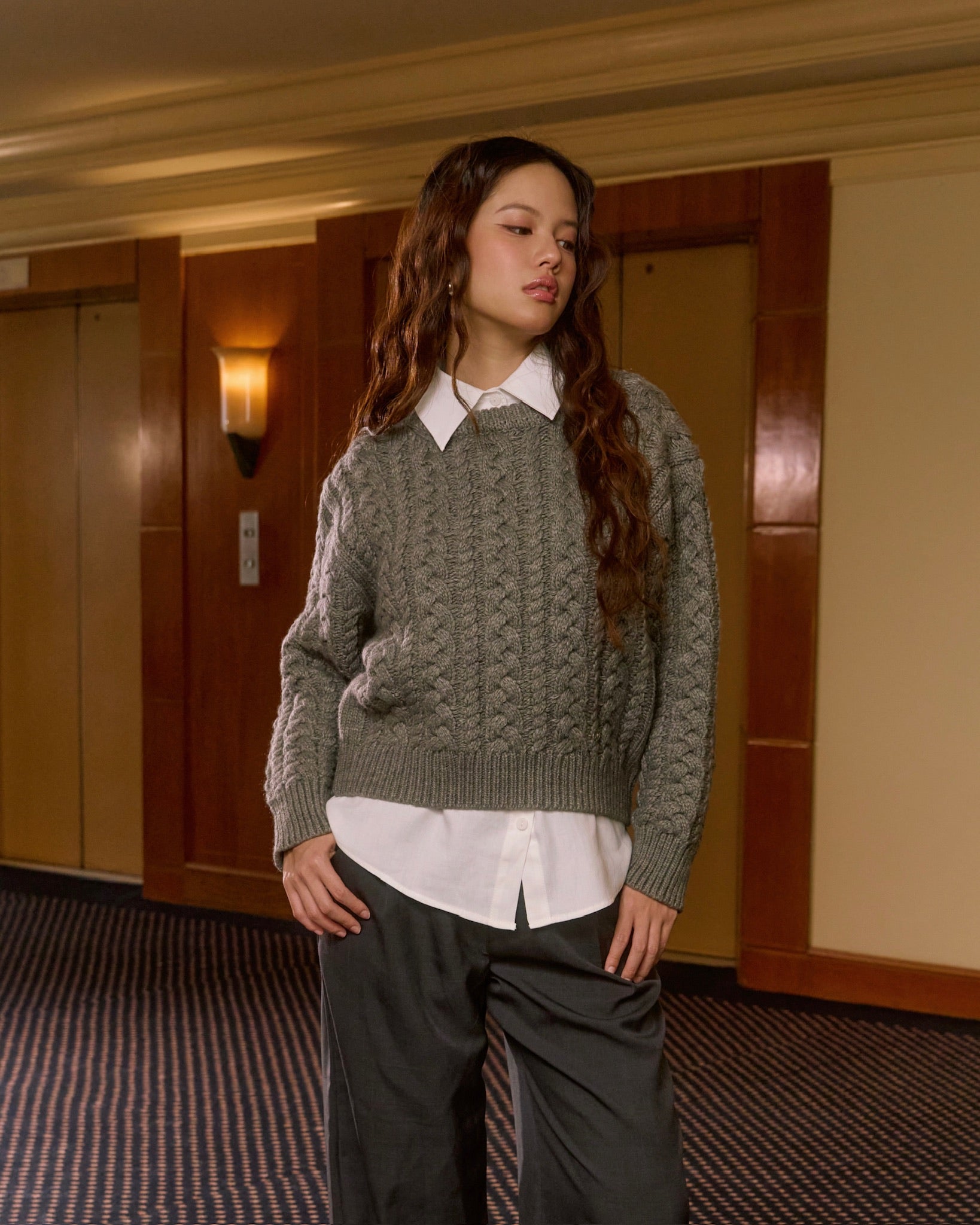 Woman wearing a gray sweater over a white shirt and dark pants in an indoor setting.