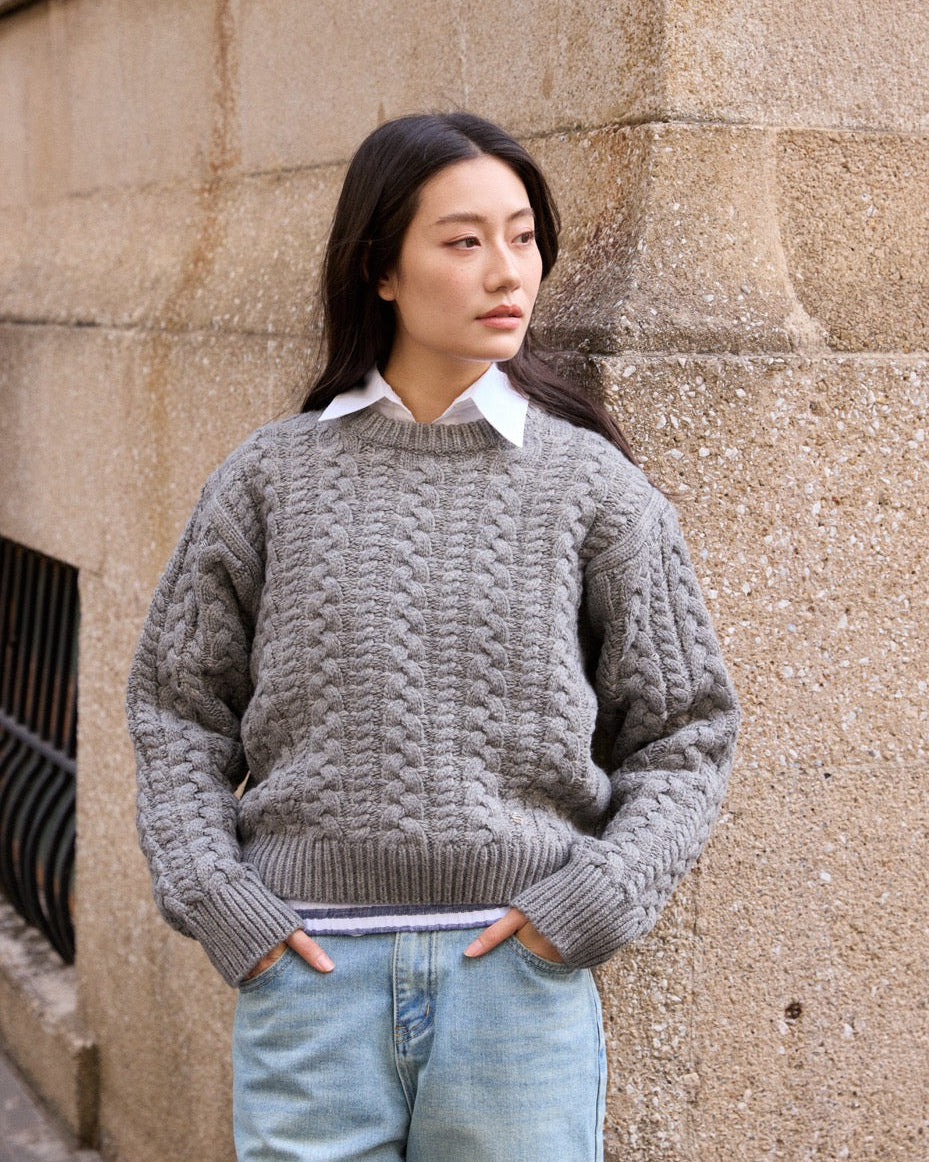 Woman wearing a gray cable knit sweater and light blue jeans against a stone wall.