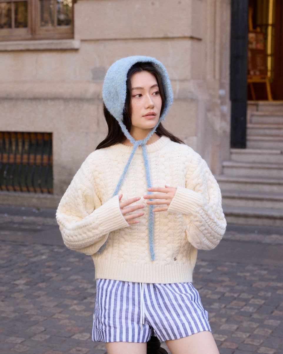 Woman wearing a cream sweater and blue striped shorts with a blue knitted bonnet outdoors.