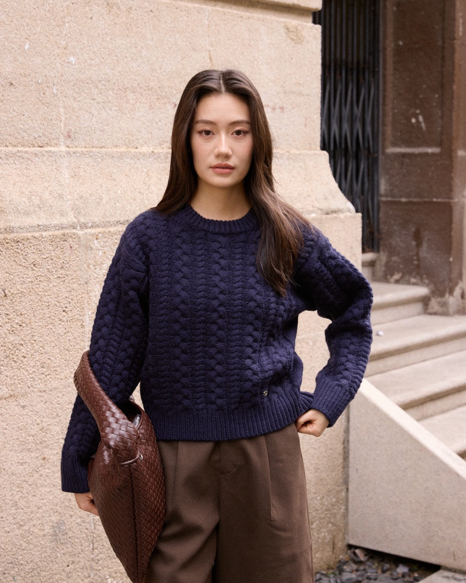 Woman wearing a dark blue sweater and brown pants standing against a stone wall.