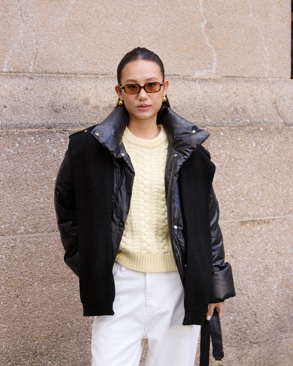 woman wearing a black coat, yellow sweater, and white pants against a beige wall.