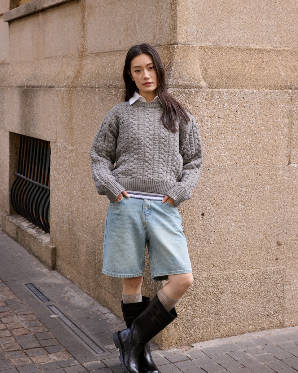Woman wearing a gray sweater, light blue shorts, and black boots against a beige wall.