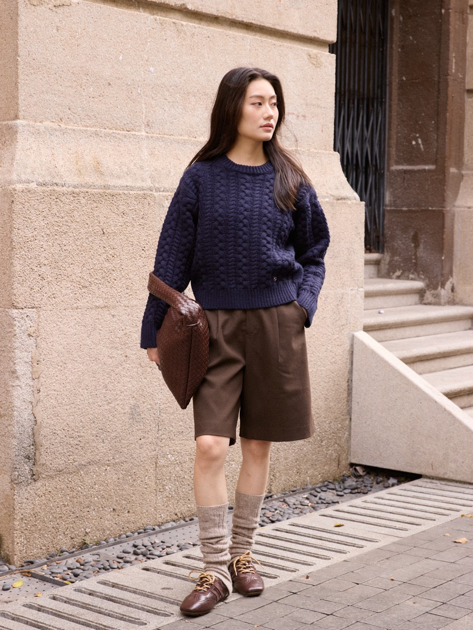 Woman wearing a navy sweater and brown shorts standing against a stone wall.