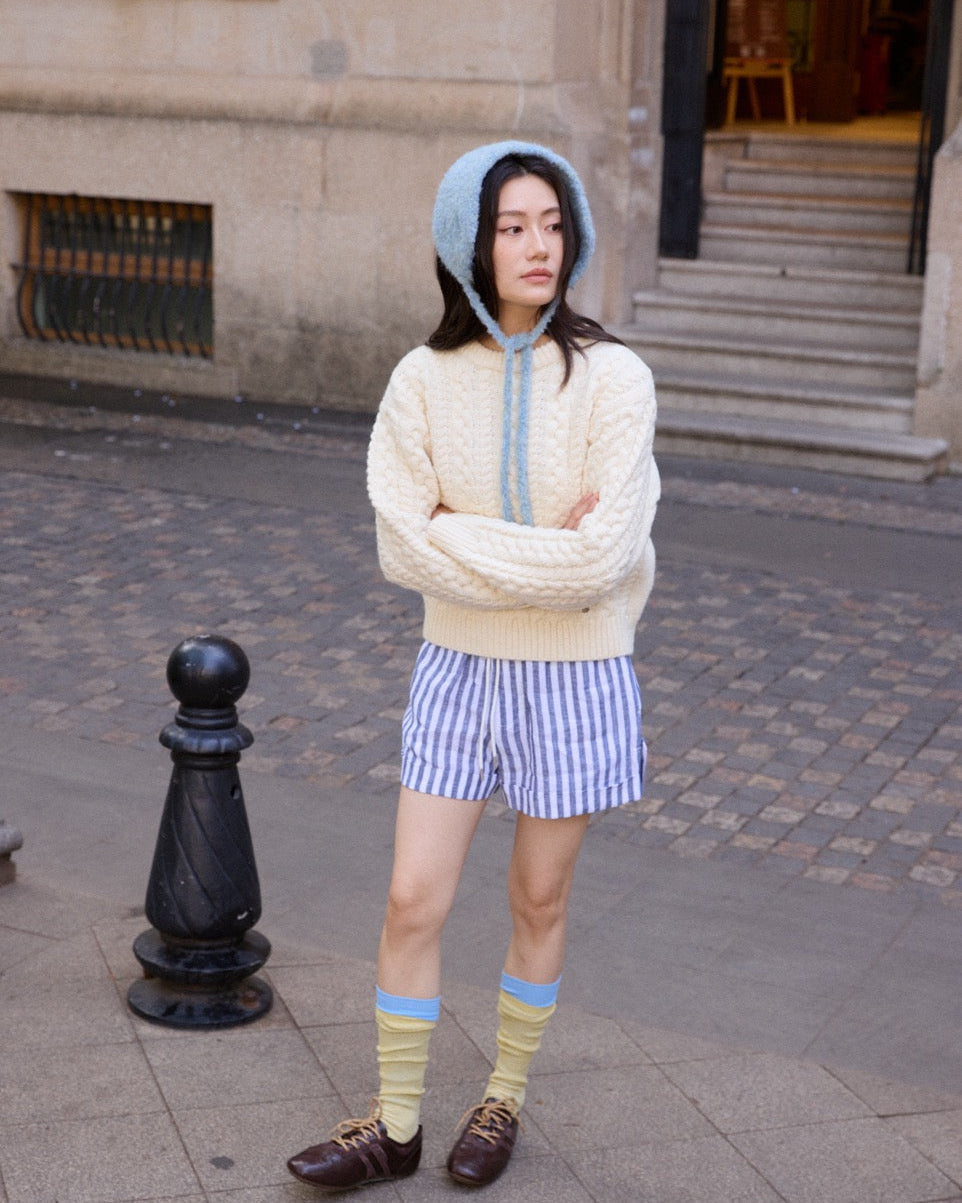 woman wearing a light-colored sweater, striped shorts, and a hooded garment in an urban setting.