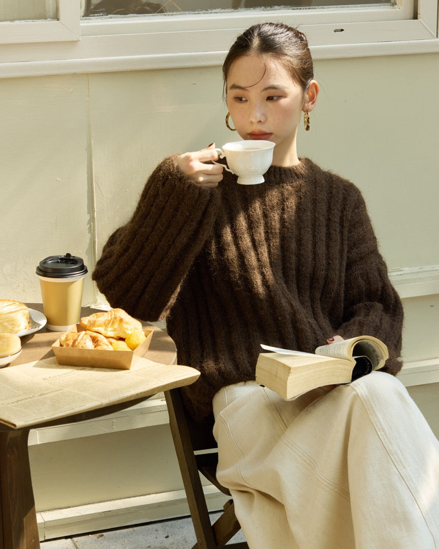 Woman sitting at a table drinking from a cup with a book and food items around her.