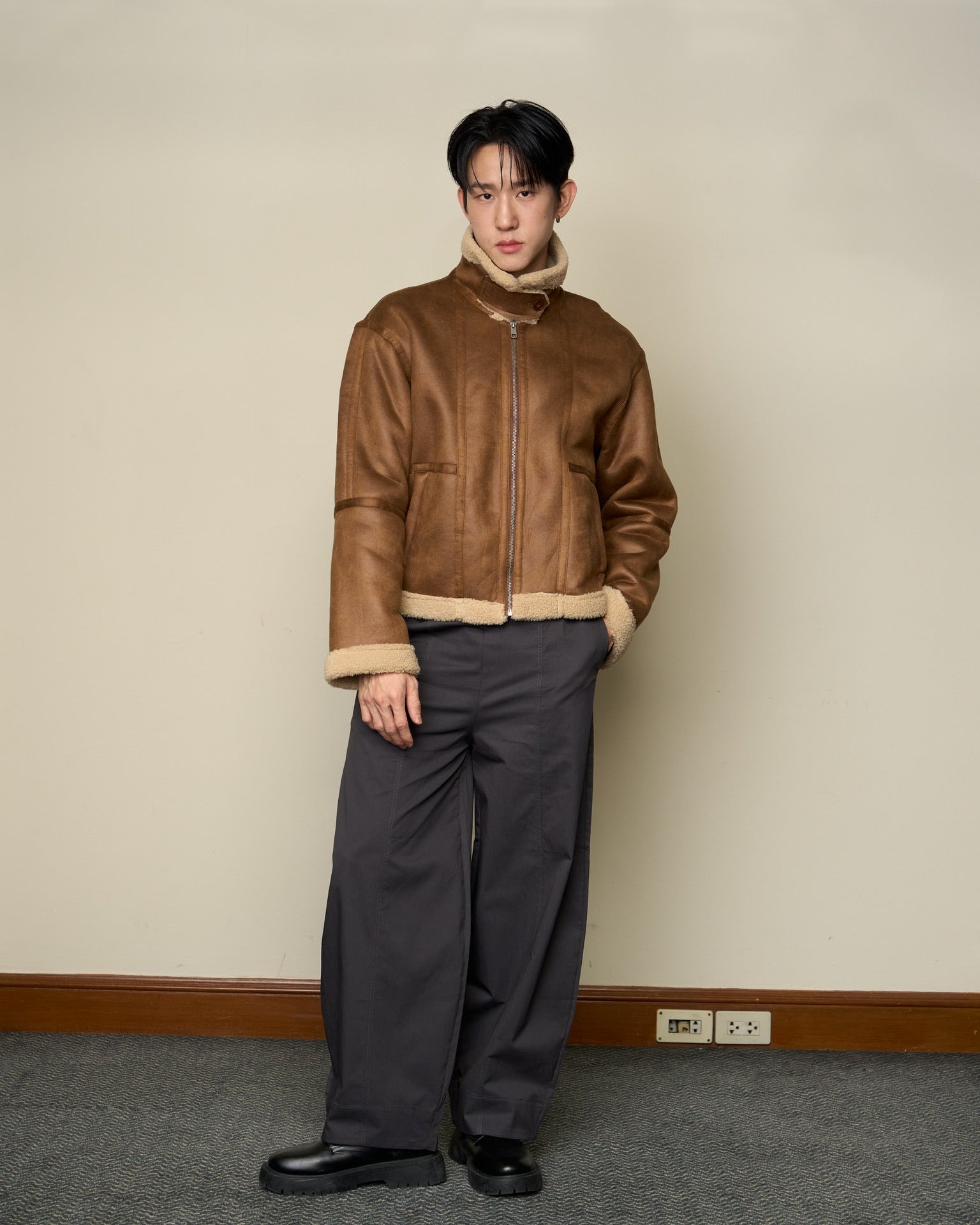 man wearing a brown leather jacket and gray pants standing against a plain wall.