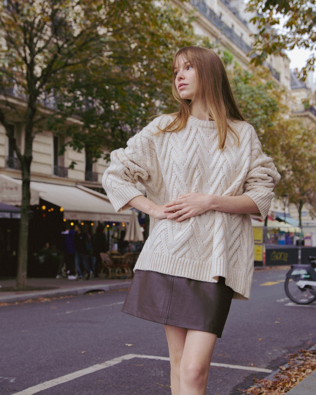 Woman wearing a cream sweater and black skirt standing on a street with trees and buildings in the background.