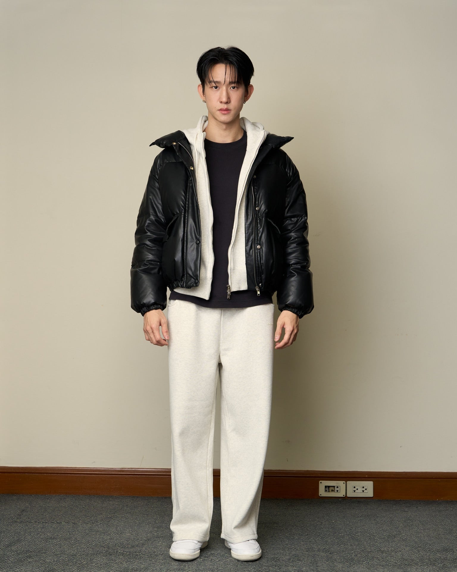 man wearing a black jacket with a white hood, black shirt, and white pants standing against a plain wall.