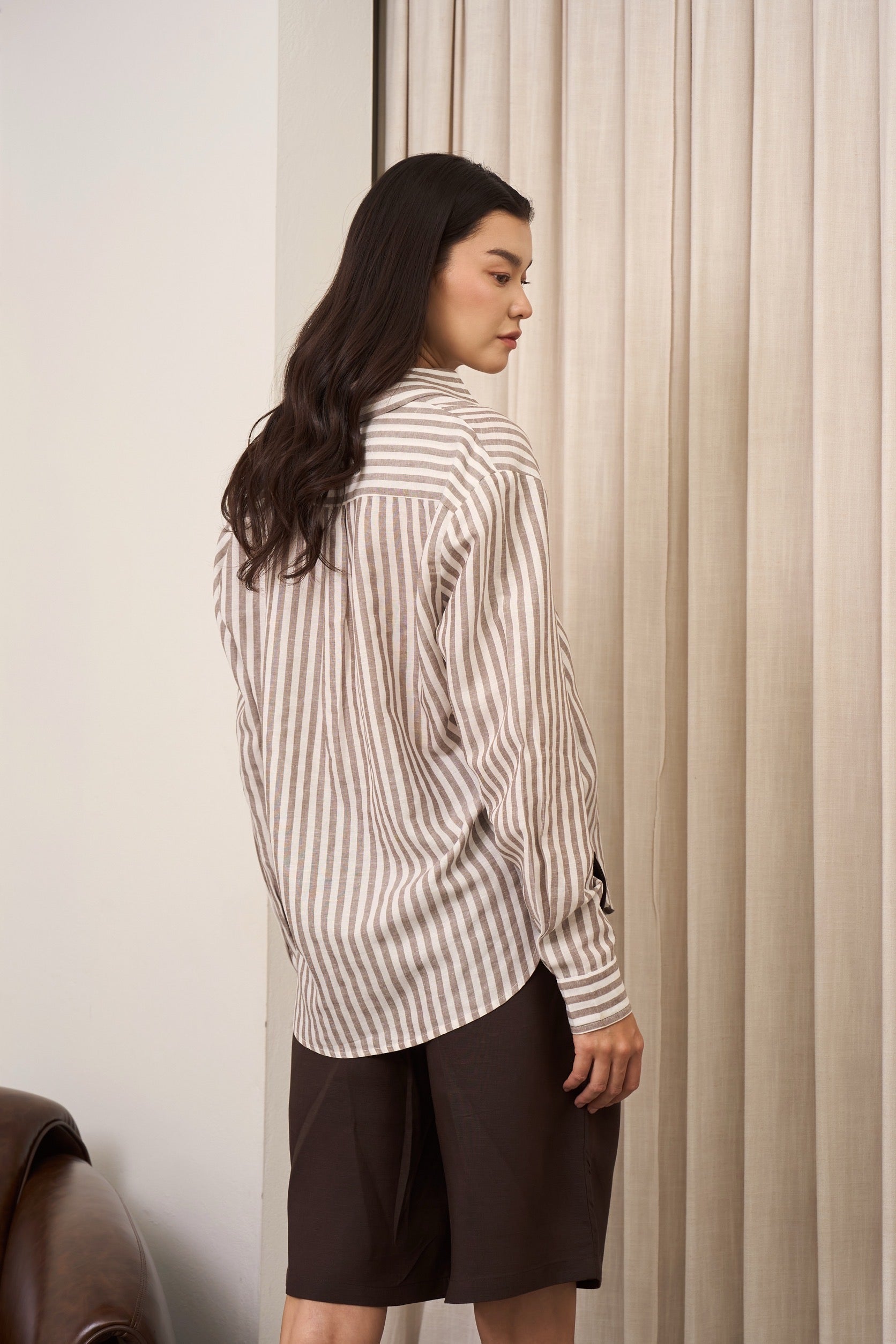 Back view of a woman wearing a brown striped shirt and brown Burmuda shorts standing facing to the wall with curtains
