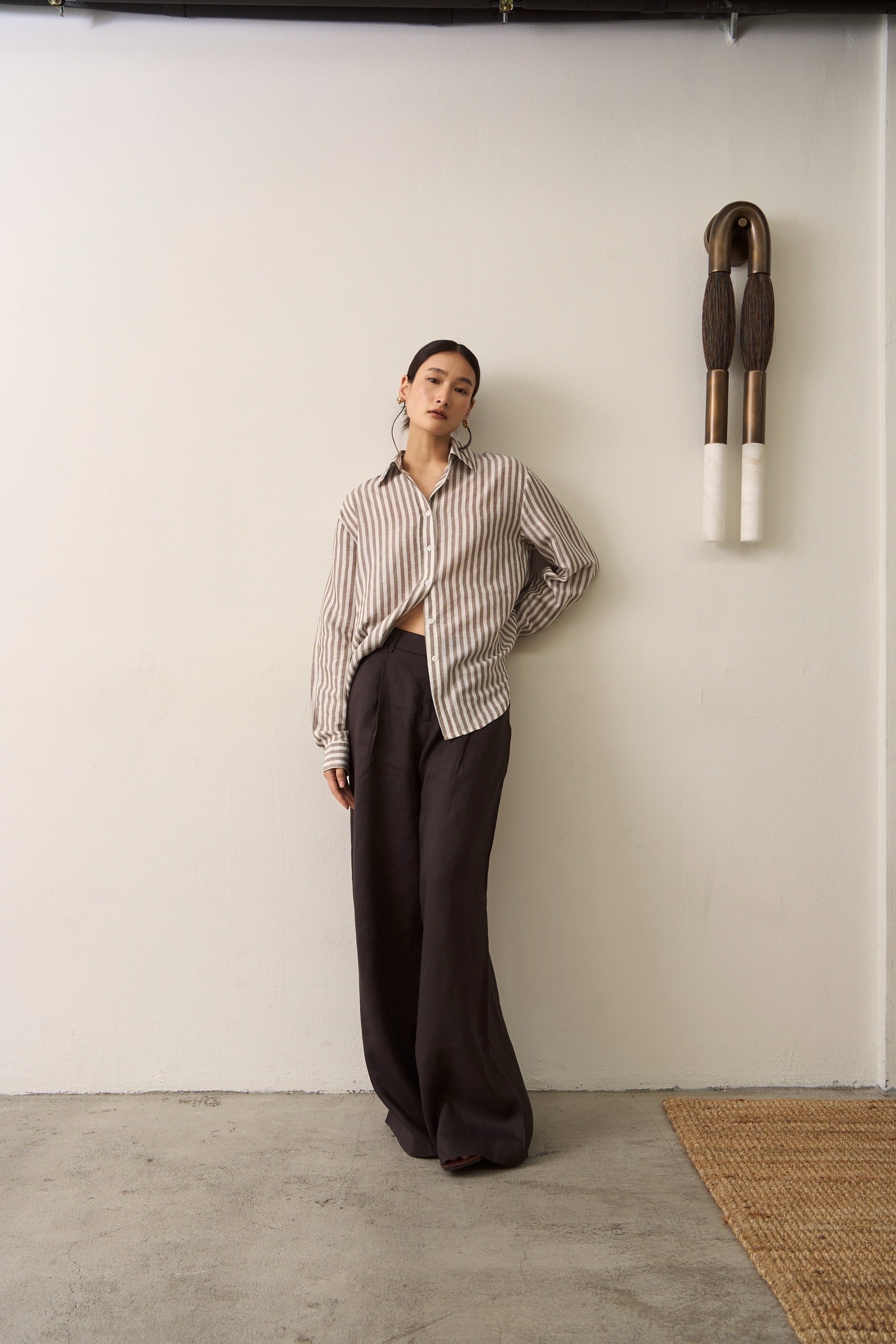 Full body view of a woman wearing a brown striped linen shirt and brown linen trousers standing against a white wall with a decorative object.