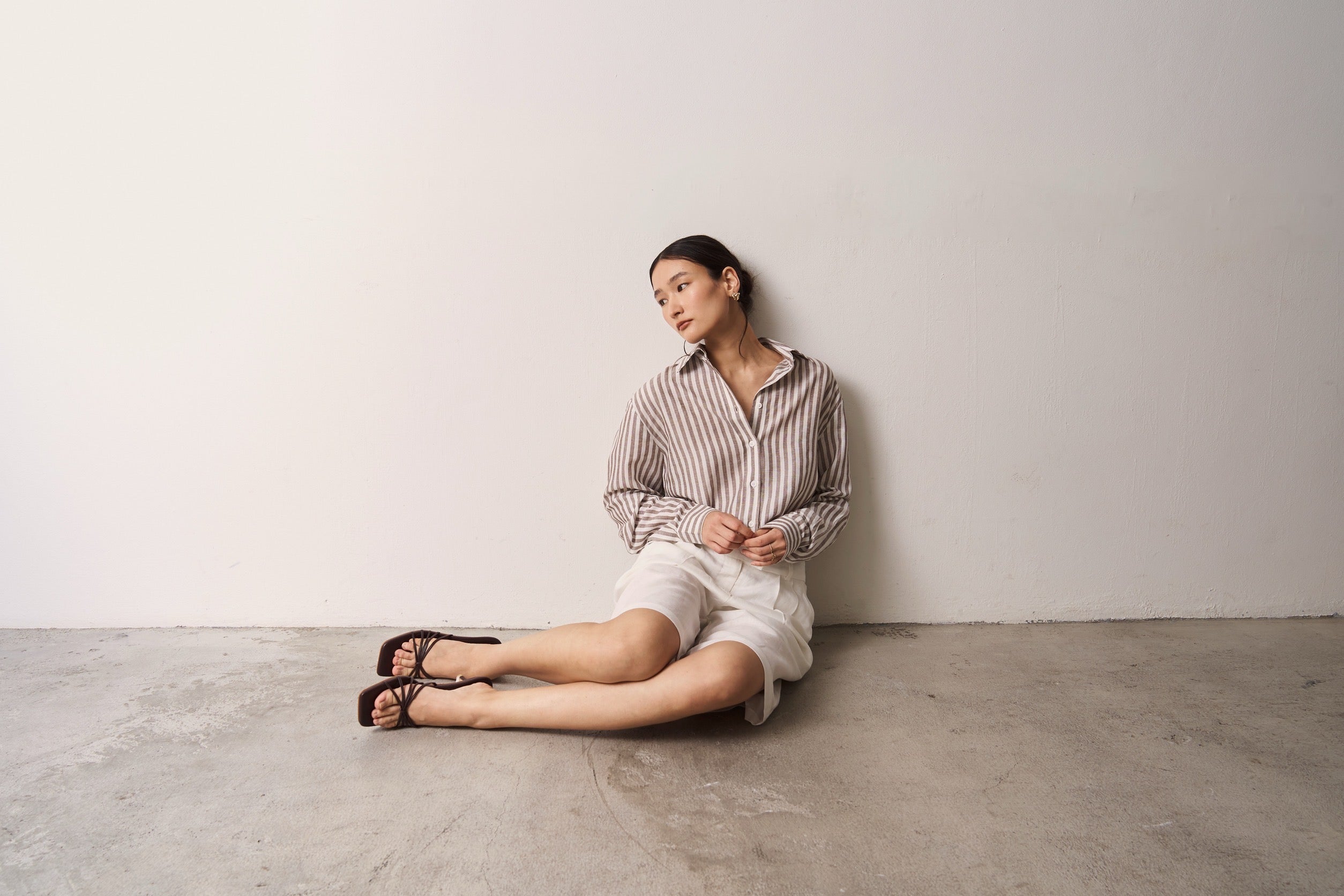 Woman wearing brown striped linen shirt and Burmuda shorts sitting against the plain wall