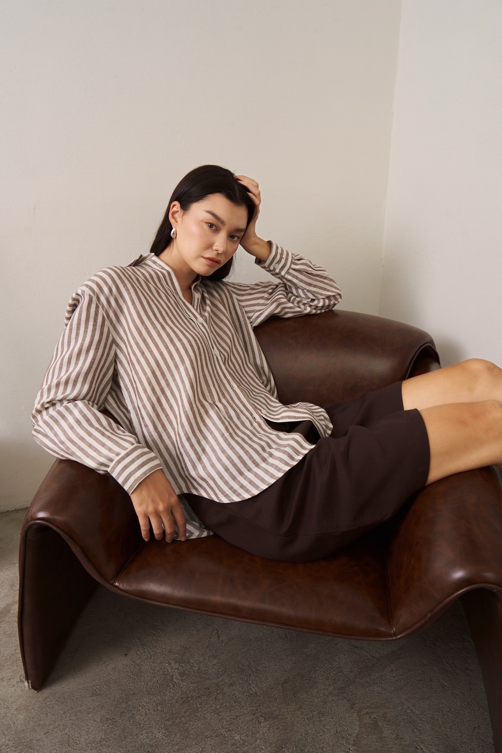 Woman sitting on a brown leather chair wearing a brown striped linen shirt and brown Burmuda shorts.