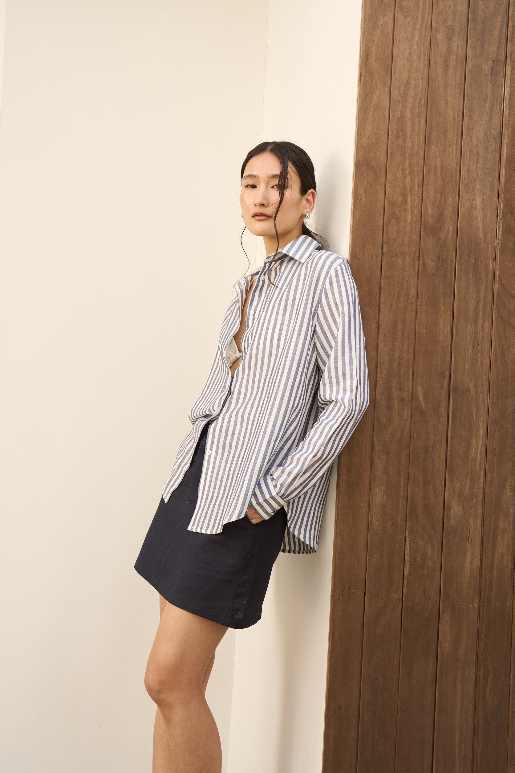 Woman wearing a long navy striped linen shirt and navy skirt against a wall.