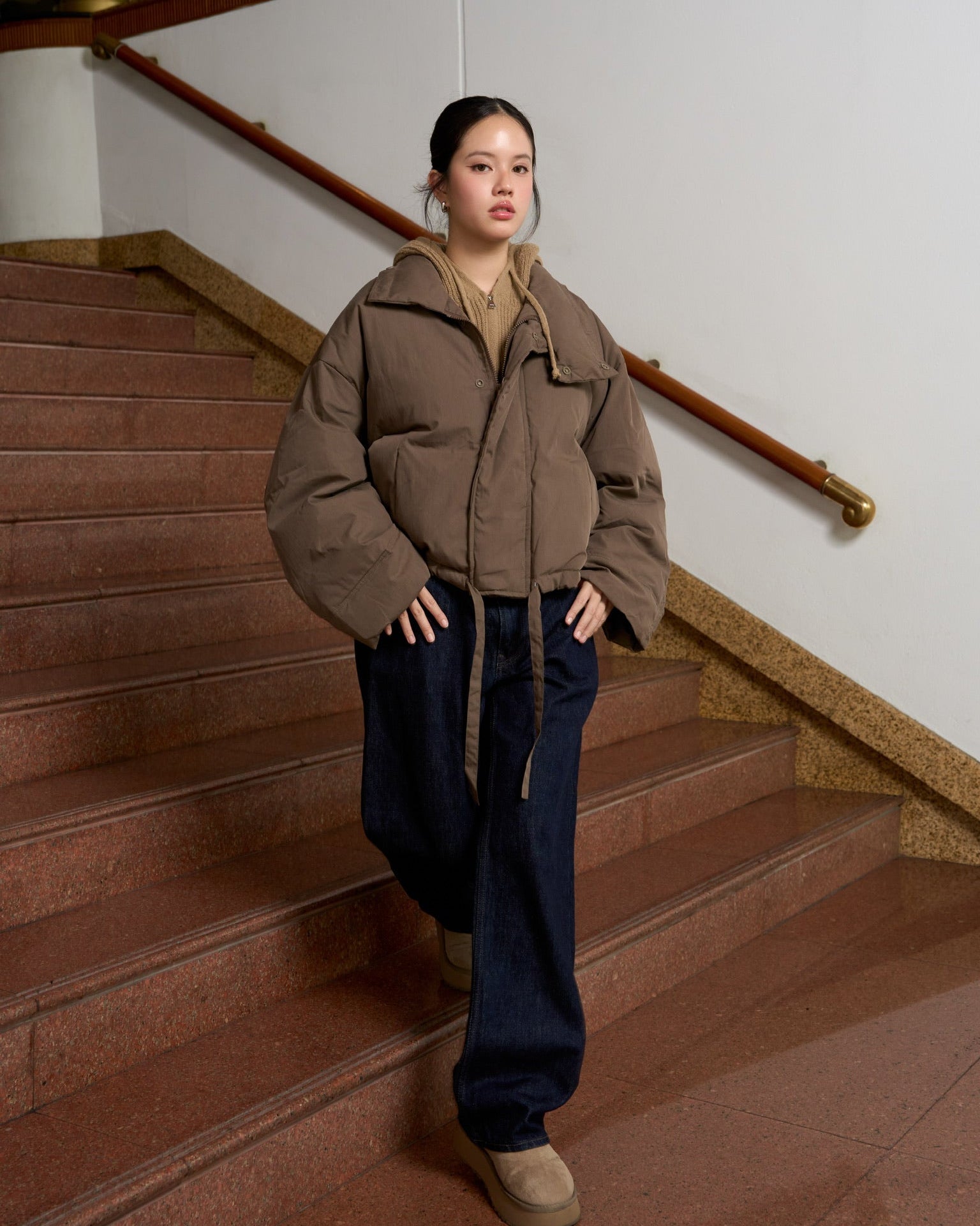 woman wearing a brown puffer jacket and dark pants standing on a staircase.