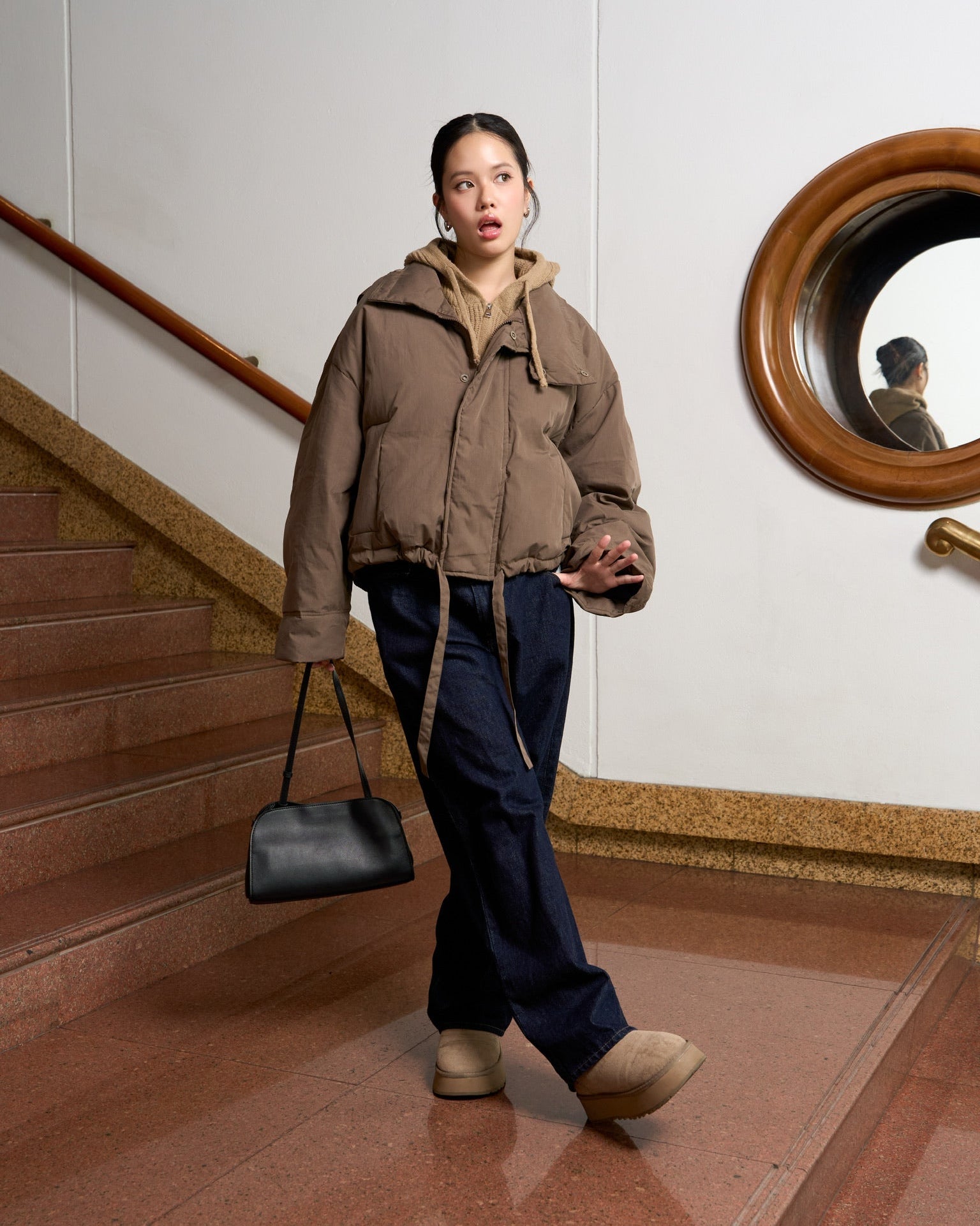 Woman in a brown jacket and dark pants standing on a staircase with a mirror in the background.