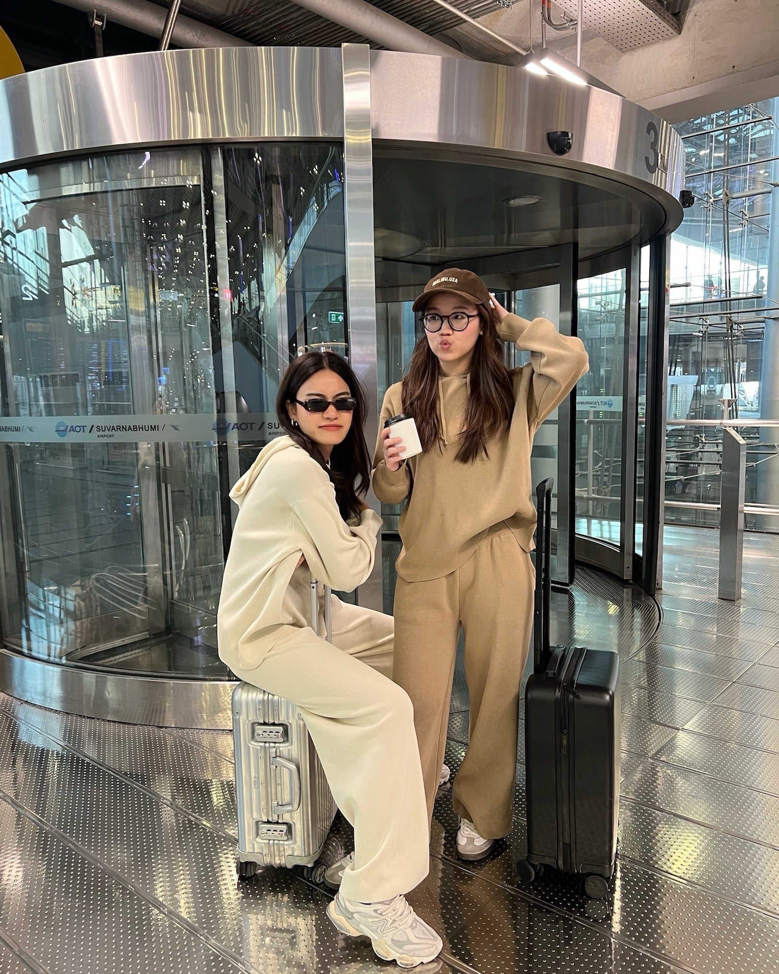 Two women in matching outfits sitting on a large suitcase in an airport.