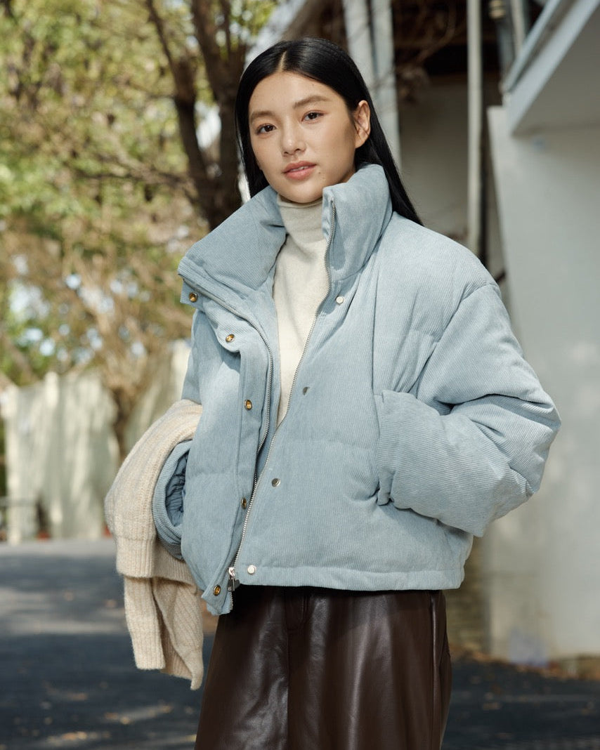 woman wearing a light blue puffer jacket outdoors with trees and a building in the background.