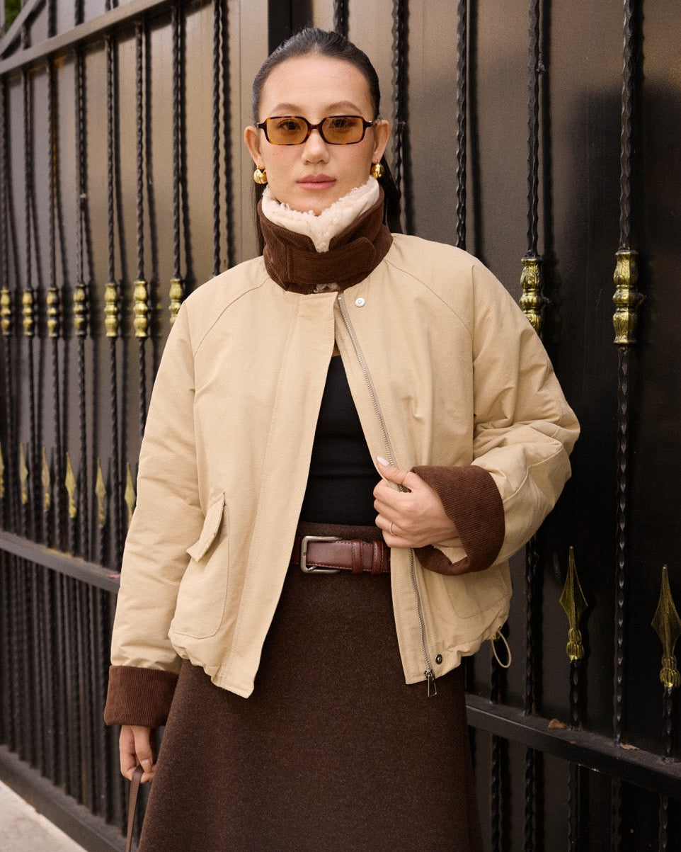 woman wearing a beige jacket with brown accents in front of a dark metal gate.