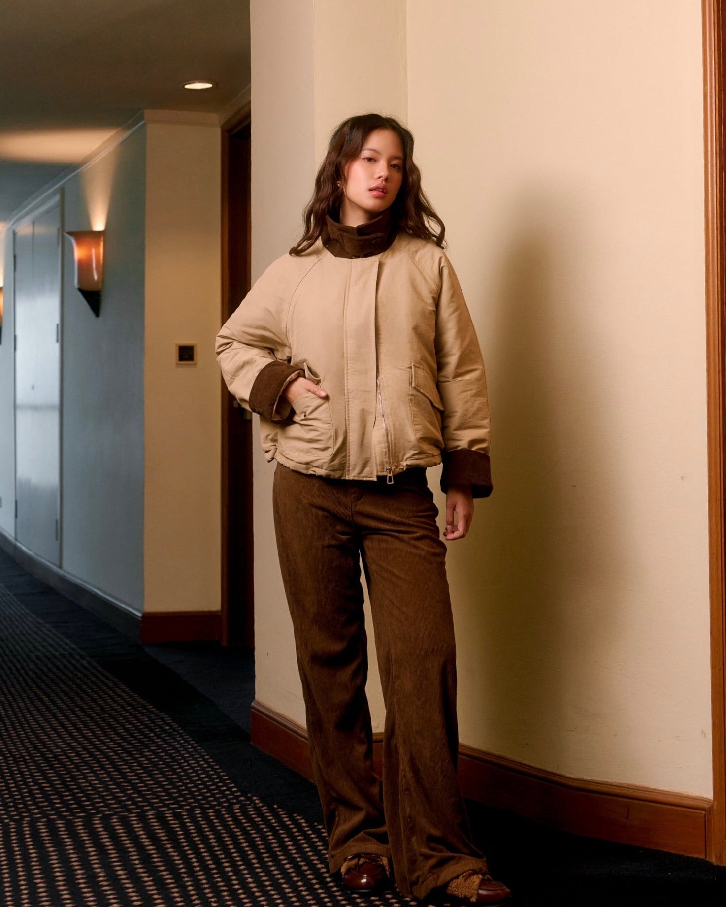 Woman standing in a hallway wearing a beige jacket and brown pants.