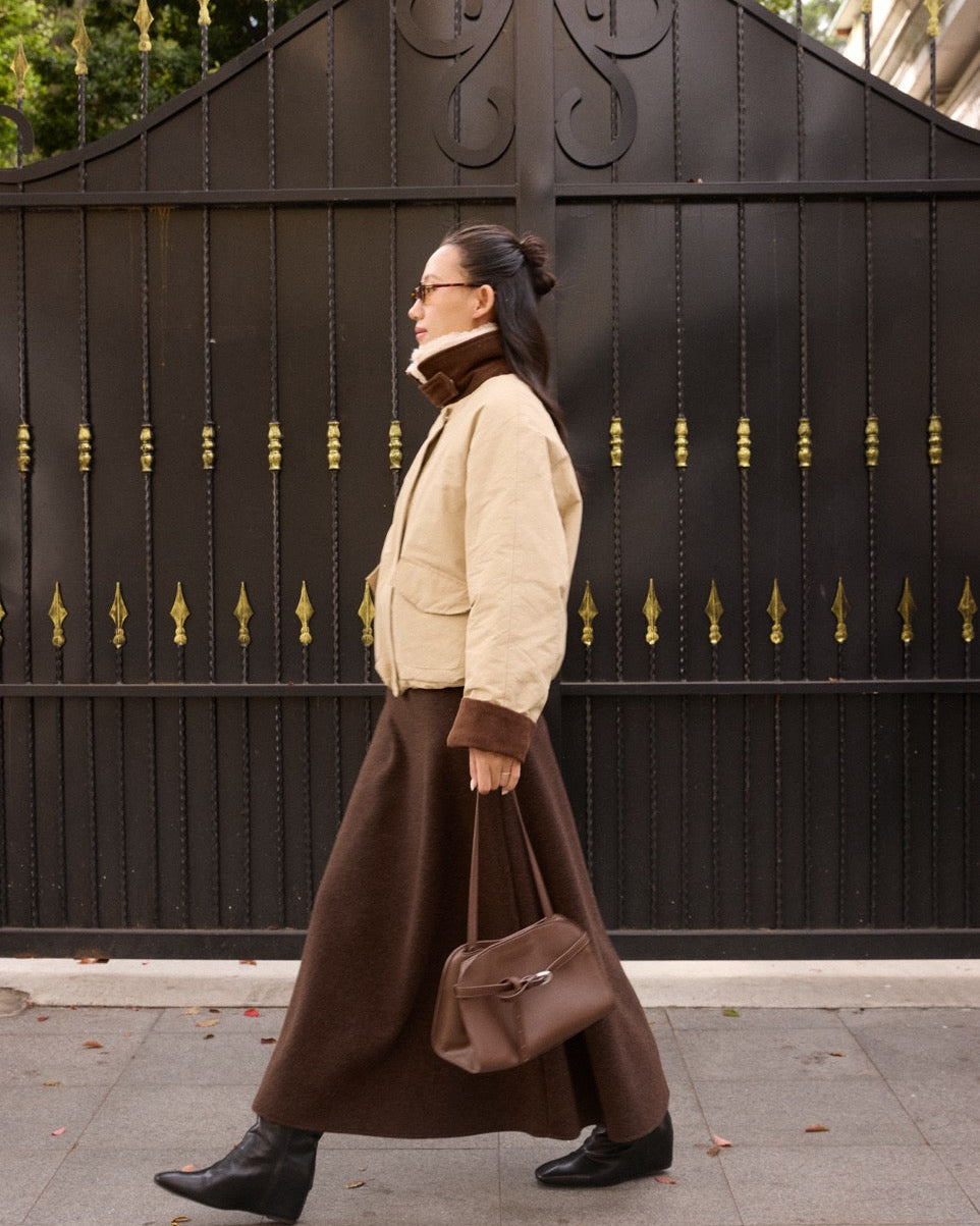 woman walking past a decorative black gate wearing a beige coat and brown skirt.