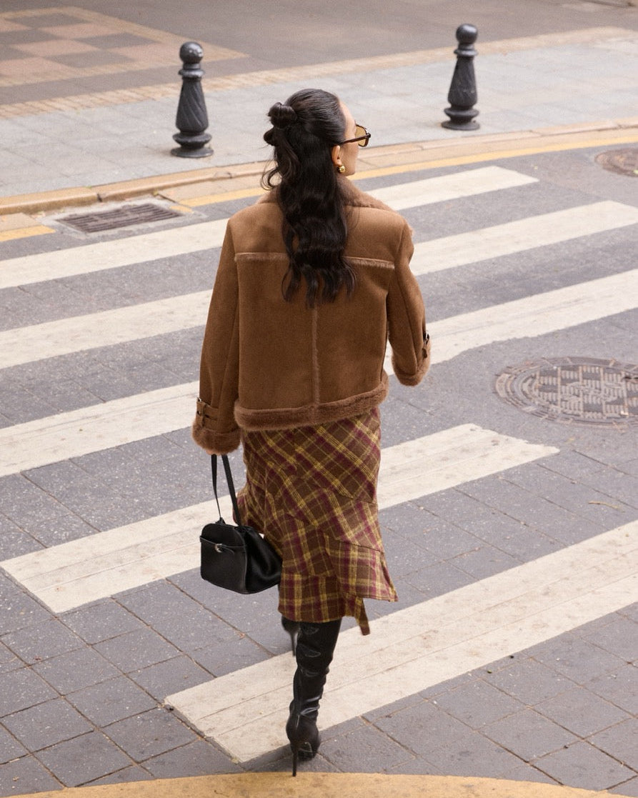 Woman in a brown coat and plaid skirt walking on a crosswalk.