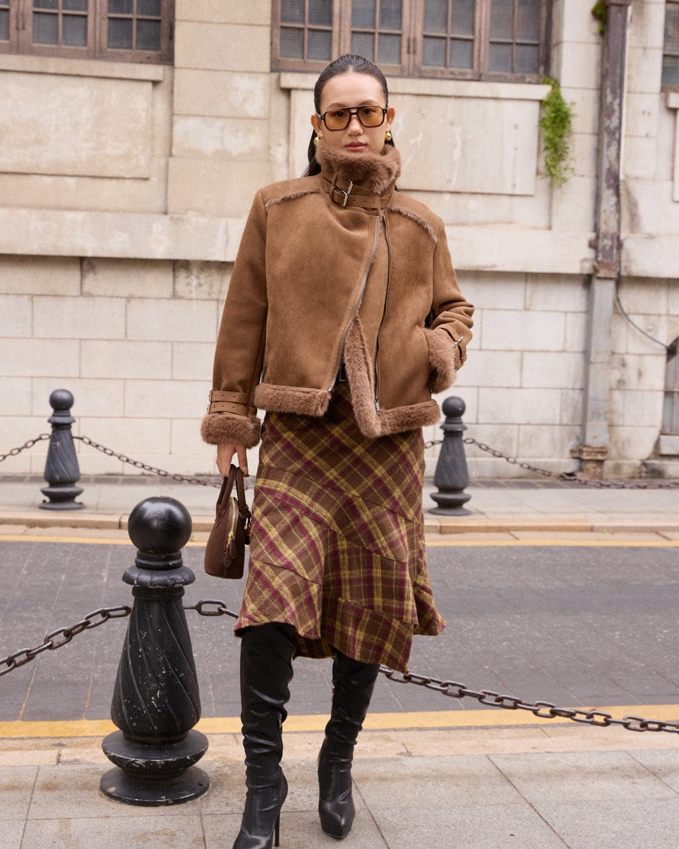 Woman wearing a brown coat and plaid skirt standing on a street.