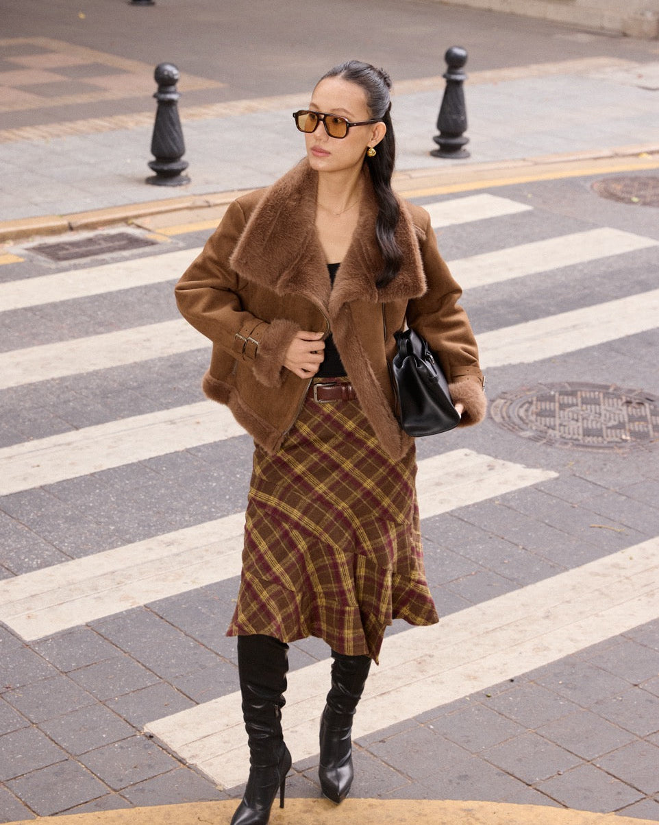 Woman in a brown coat and plaid skirt crossing a street.