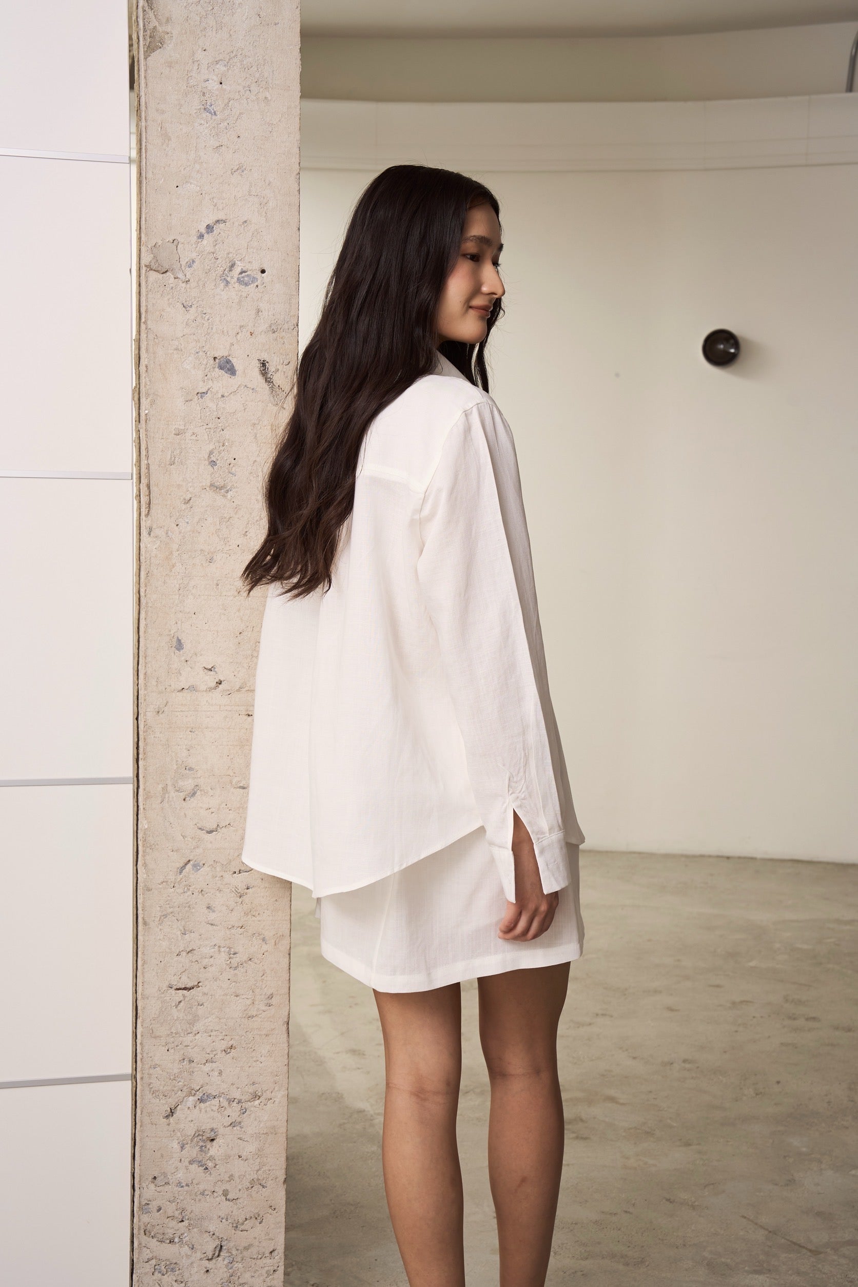 Woman wearing a white linen shirt and skirt outfit standing against a neutral wall.