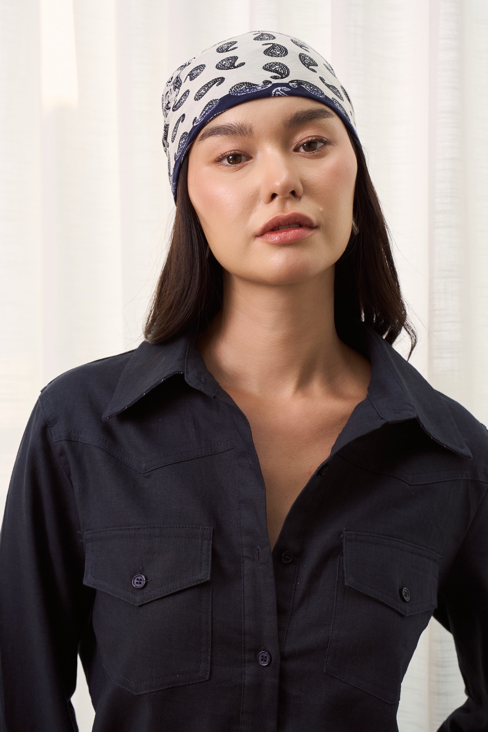 Woman wearing a dark navy linen shirt with double pockets and patterned headscarf against a white curtain background