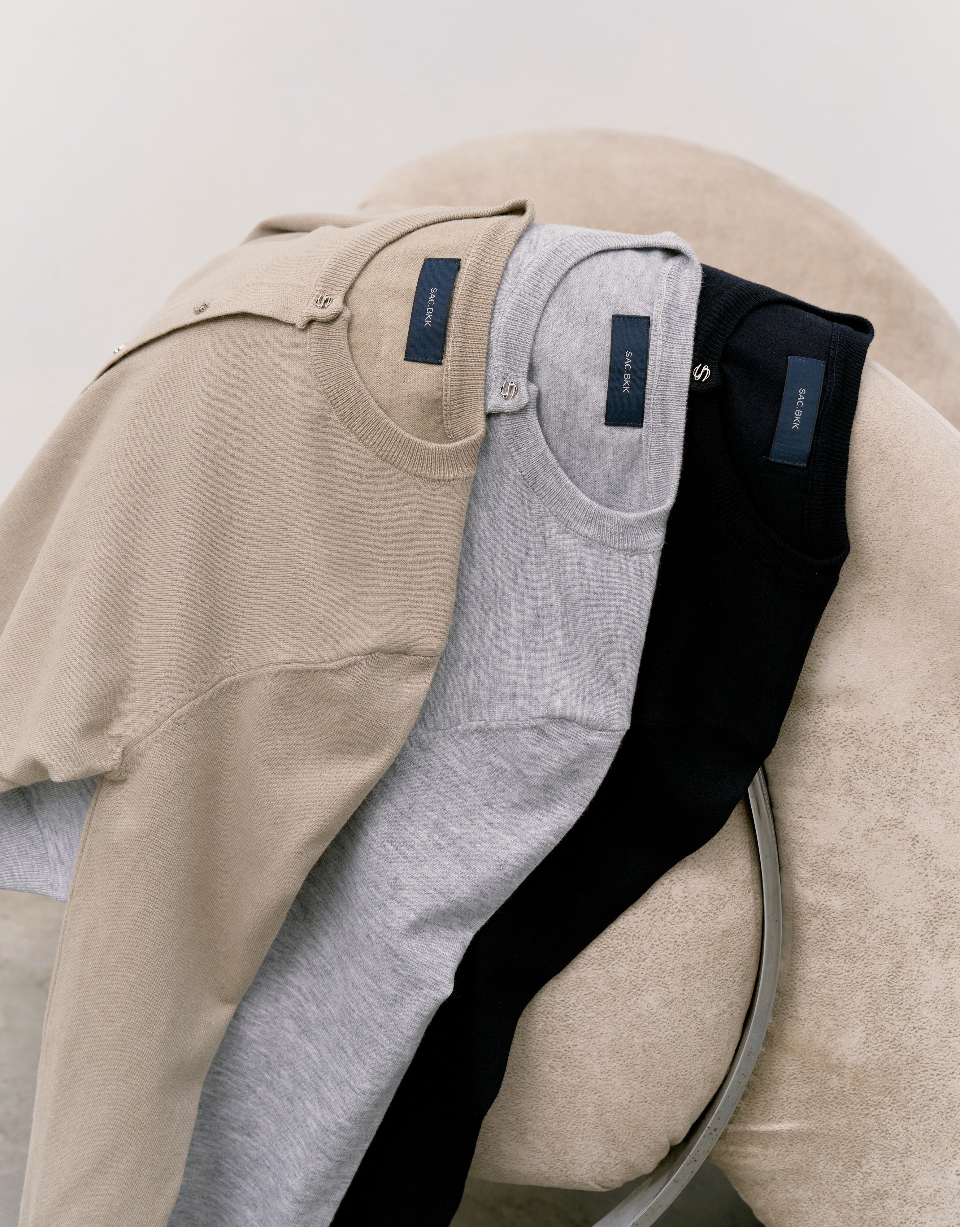 Three pairs of jeans in beige, gray, and black on a beige cushion against a white background.