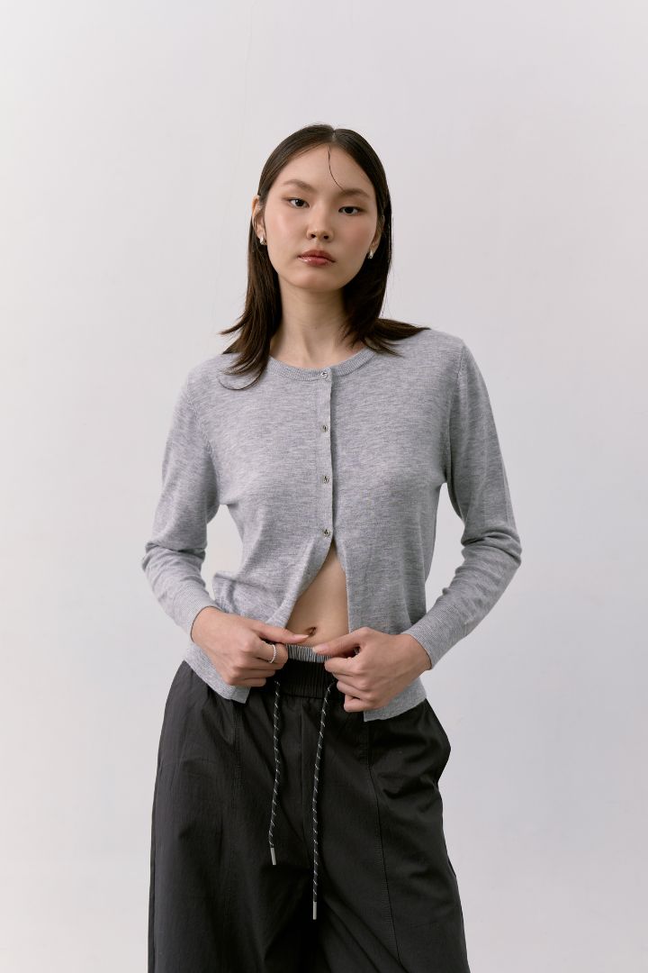 Woman wearing a light gray cardigan and black barrel pants, standing against a plain light gray studio background.