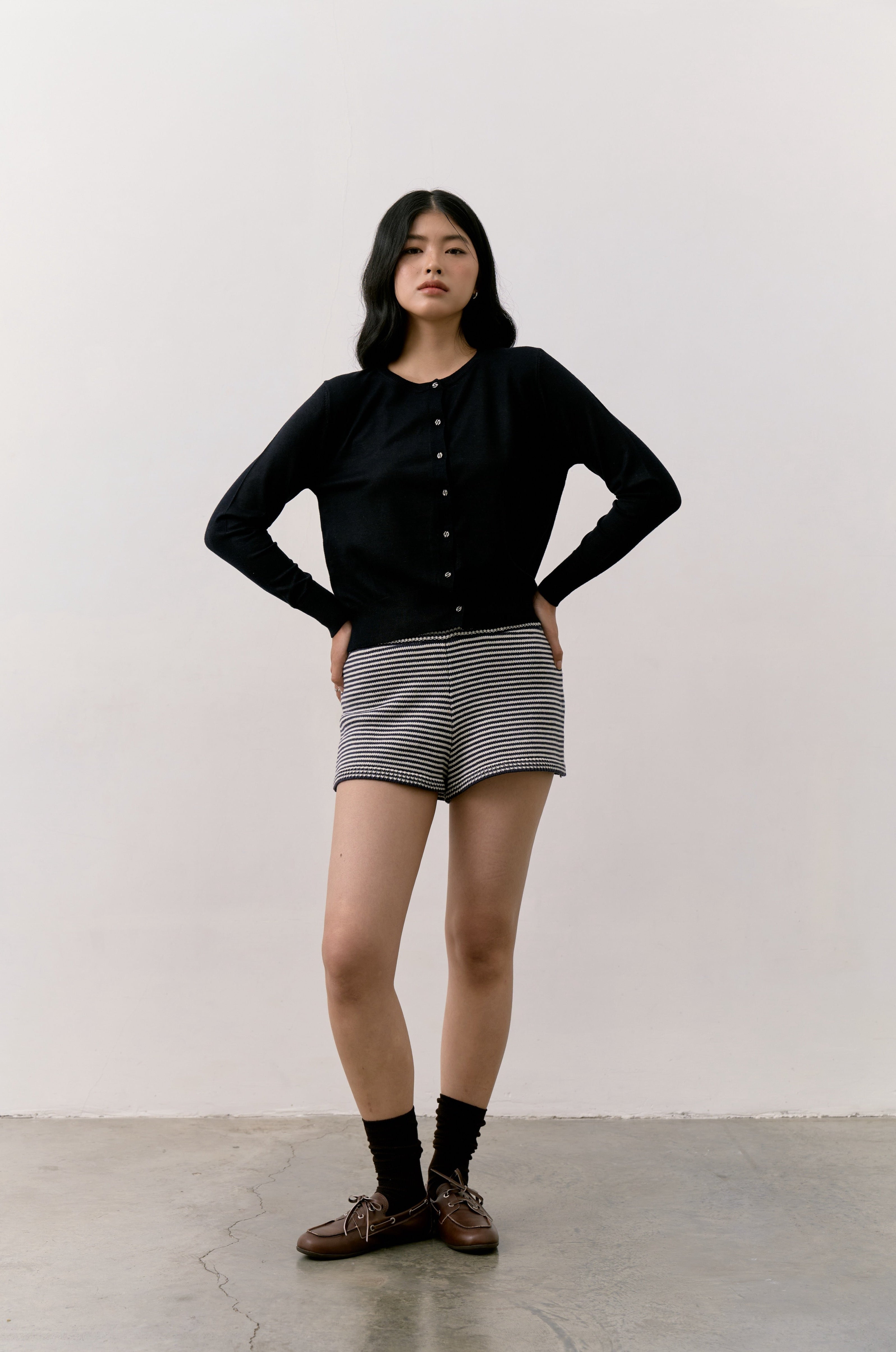 Woman wearing a black Double S Cardigan, striped shorts, and brown shoes against a white background.