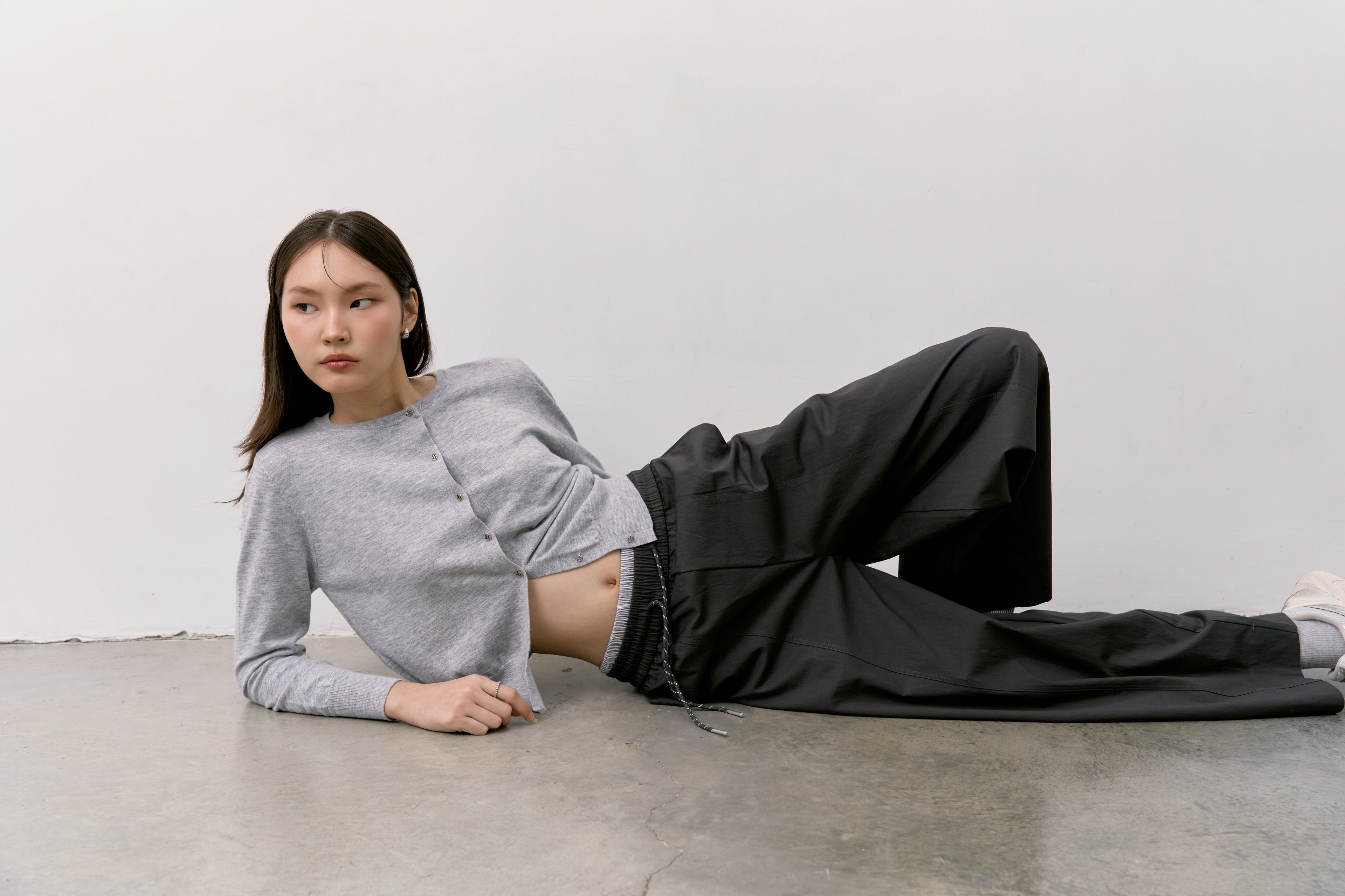 Person wearing a gray Double S Cardigan and black barrel pants lying on a concrete floor against a white wall.