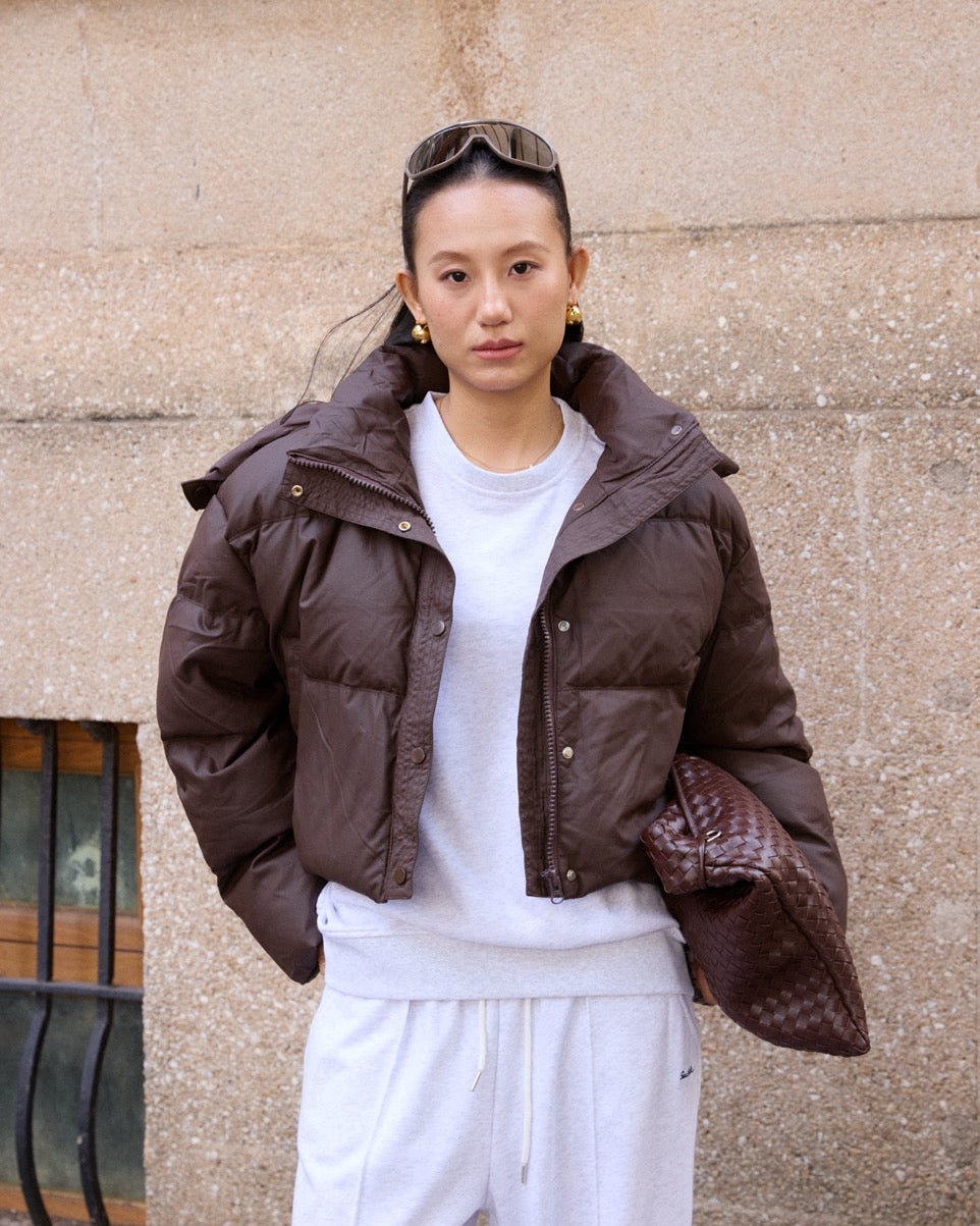 woman wearing a brown puffer jacket over a white outfit against a beige wall.