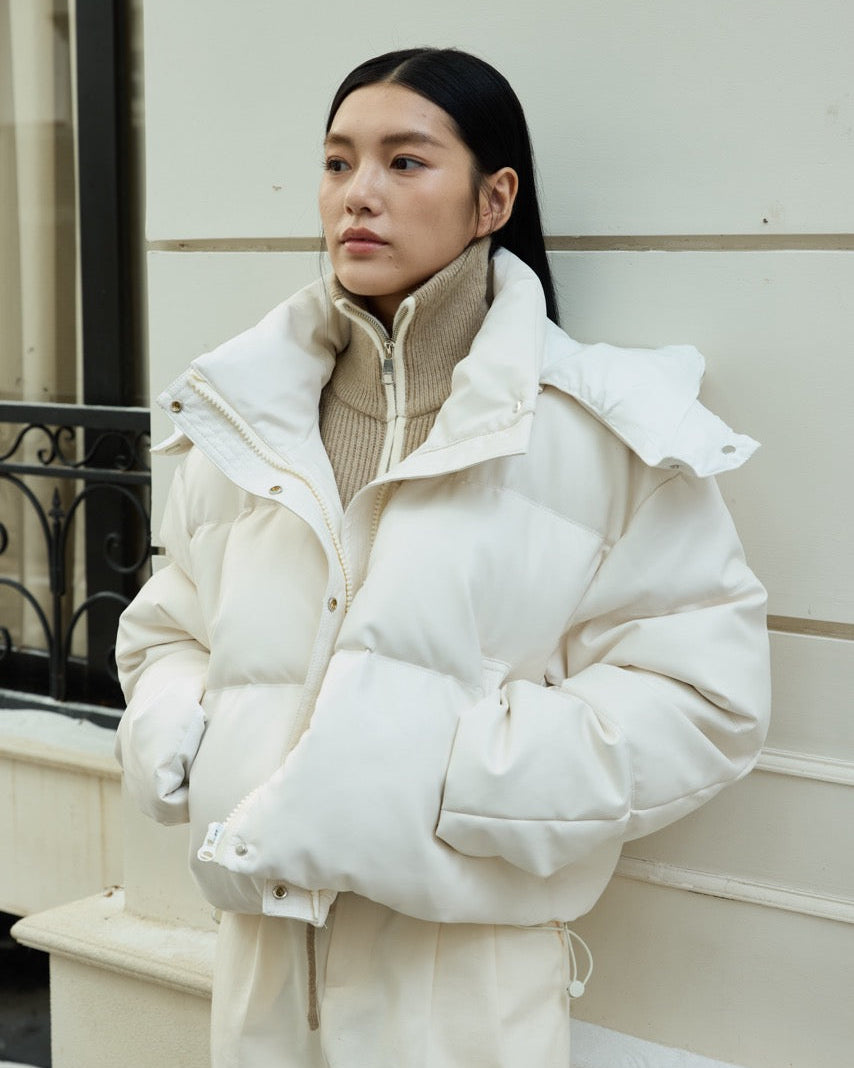 woman wearing a white puffer jacket and beige sweater against a light-colored wall.