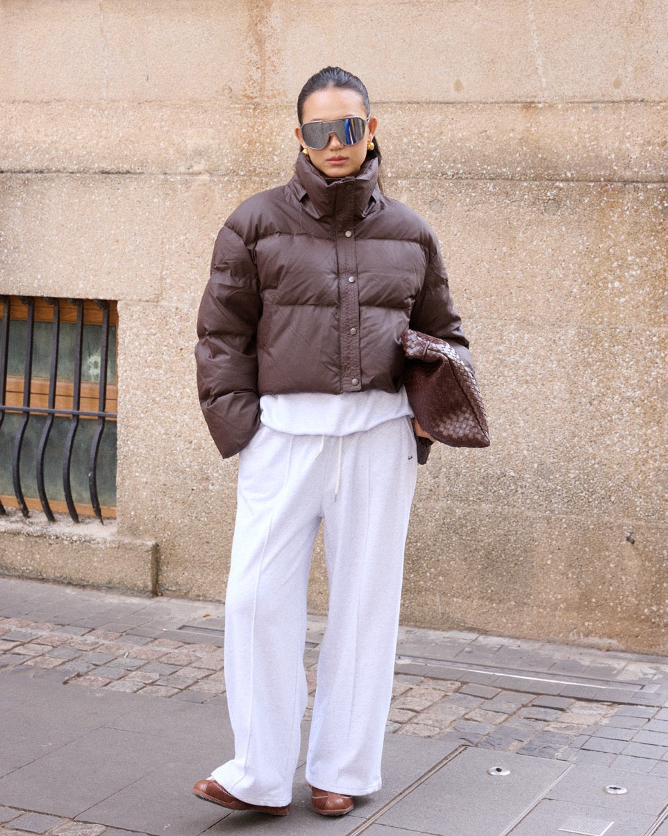 woman wearing a brown puffer jacket and white pants standing against a beige wall.