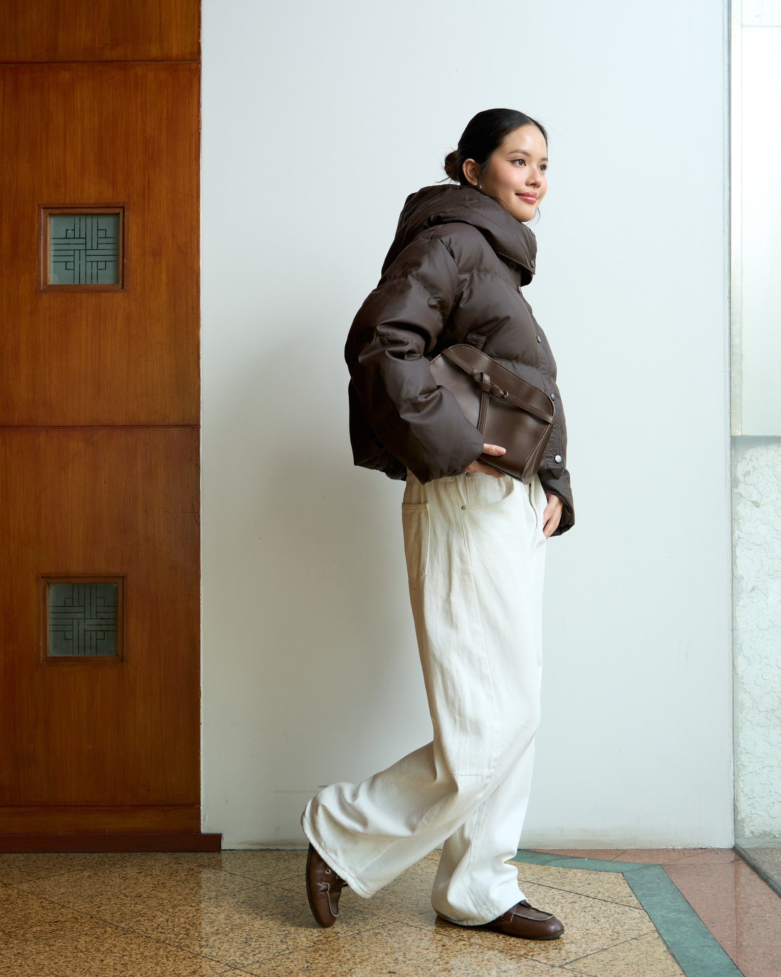 woman wearing a brown puffer jacket and white pants standing in a room with a wooden door and light-colored floor.