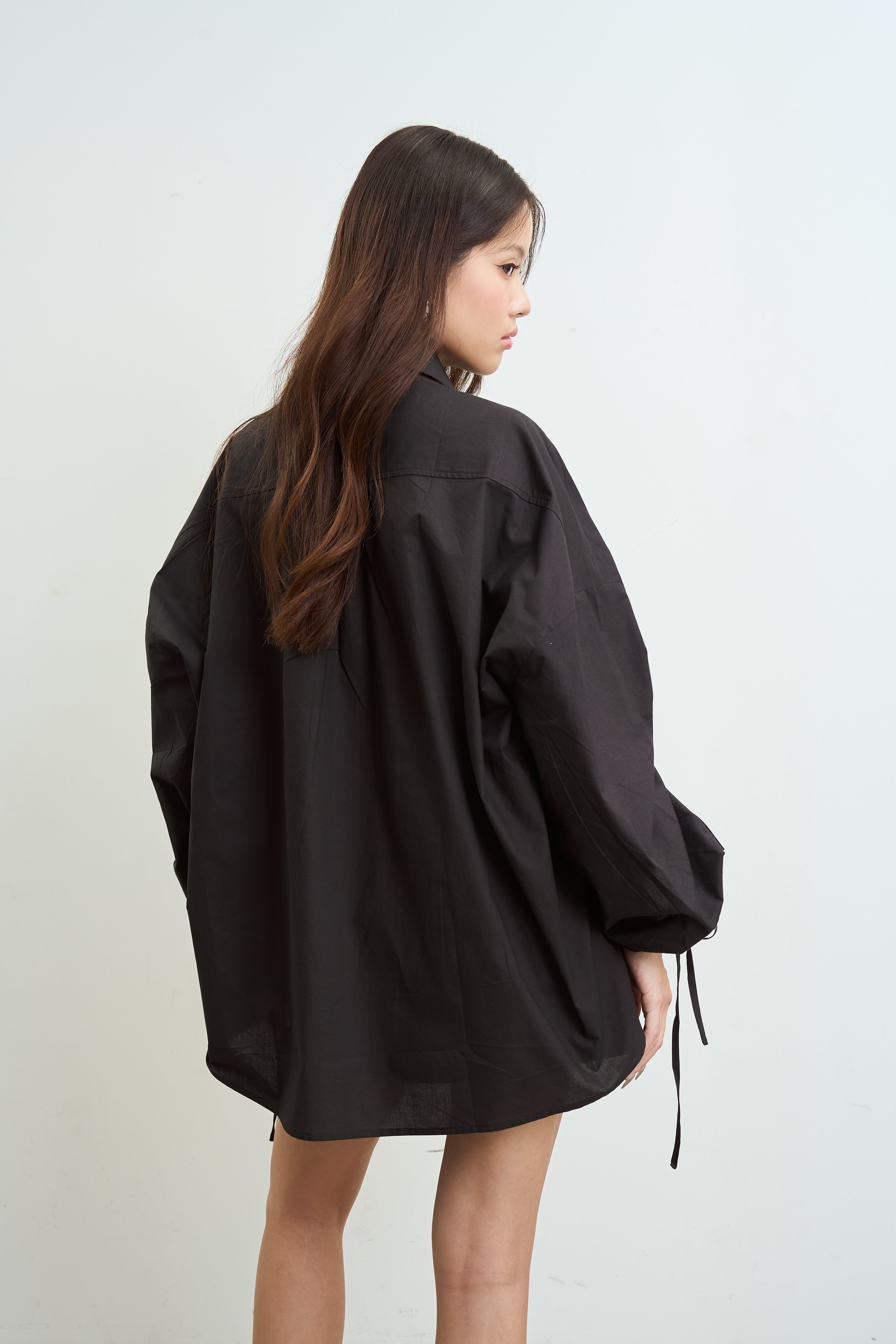 woman with long brown hair is shown from the back wearing an oversized black long-sleeve shirt with drawstrings at the cuffs, photographed against a plain light gray studio background.