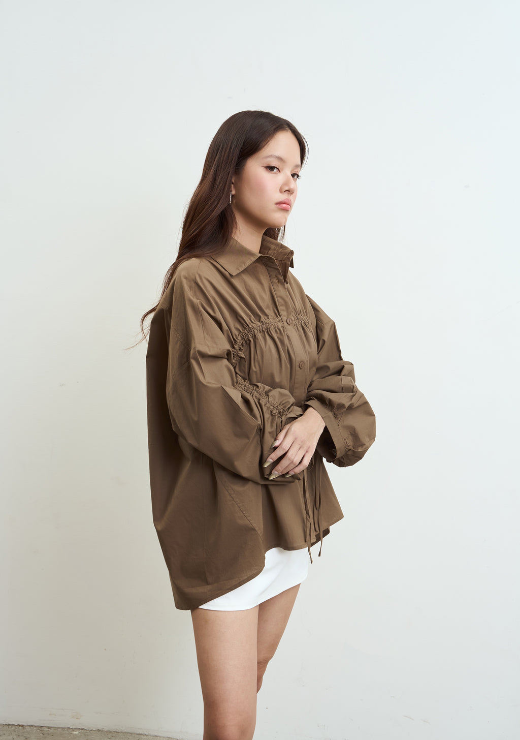 woman modeling a brown oversized long-sleeve utility-style jacket with gathered detailing and drawstrings, paired with white shorts, standing against a plain off-white wall.