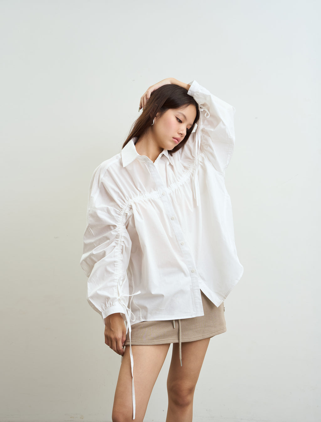 woman wearing an oversized white long-sleeve button-up shirt with gathered sleeves and drawstring ties, paired with a beige mini skirt, posing against a plain off-white background.