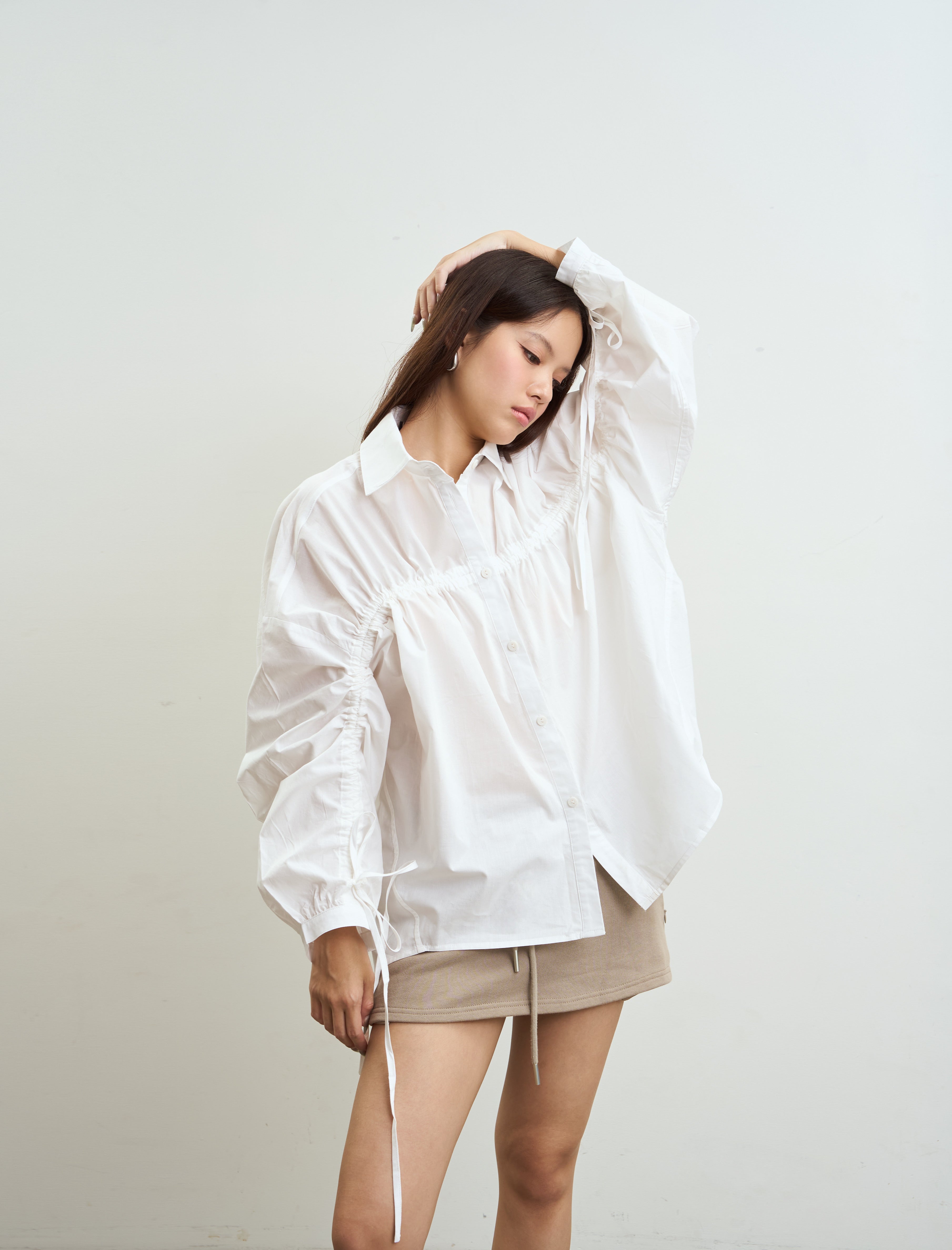 woman wearing an oversized white long-sleeve button-up shirt with gathered sleeves and drawstring ties, paired with a beige mini skirt, posing against a plain off-white background.