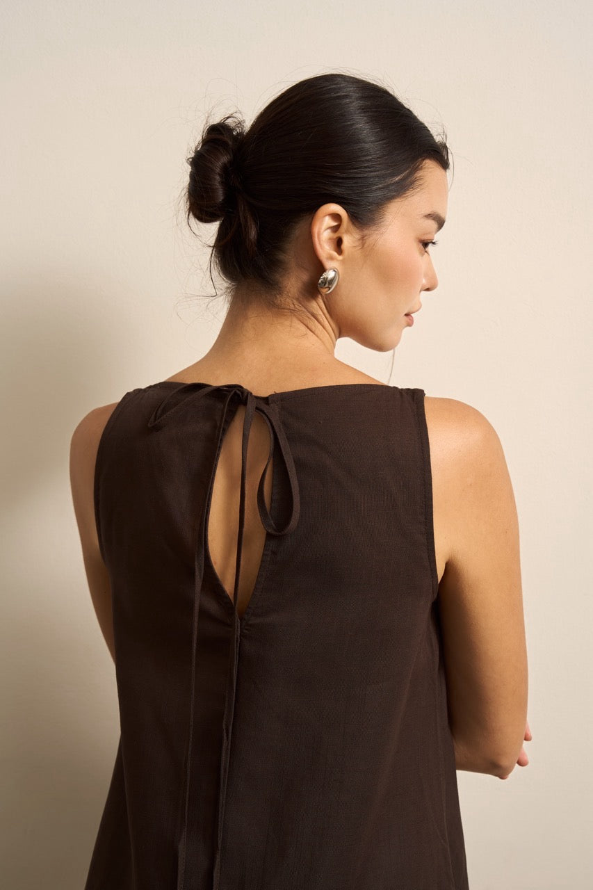 Back of a woman wearing a brown sleeveless linen dress with a bow detail on a beige background